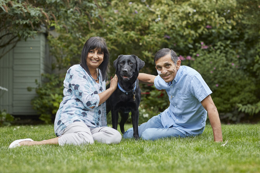 Volunteer fosterers Ron and Prisca sat on their garden lawn with black Labrador Nesta 