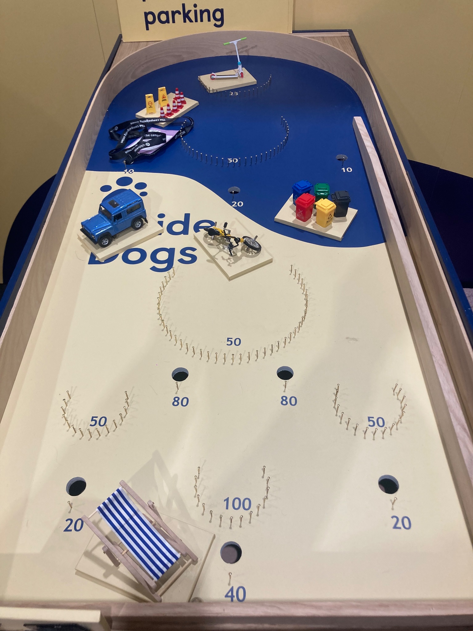 Photo of the Guide Dogs party conference pinball game, the board is yellow and blue and features the Guide Dogs logo. There are various scoring zones marked with pins and numbers indicating the potential score. Placed on the pinball board are obstacles which prevent people with a vision impairment from being able to access the world, such as pavement parking, street clutter, bins left on pavements and dockless bikes and e-scooters. 