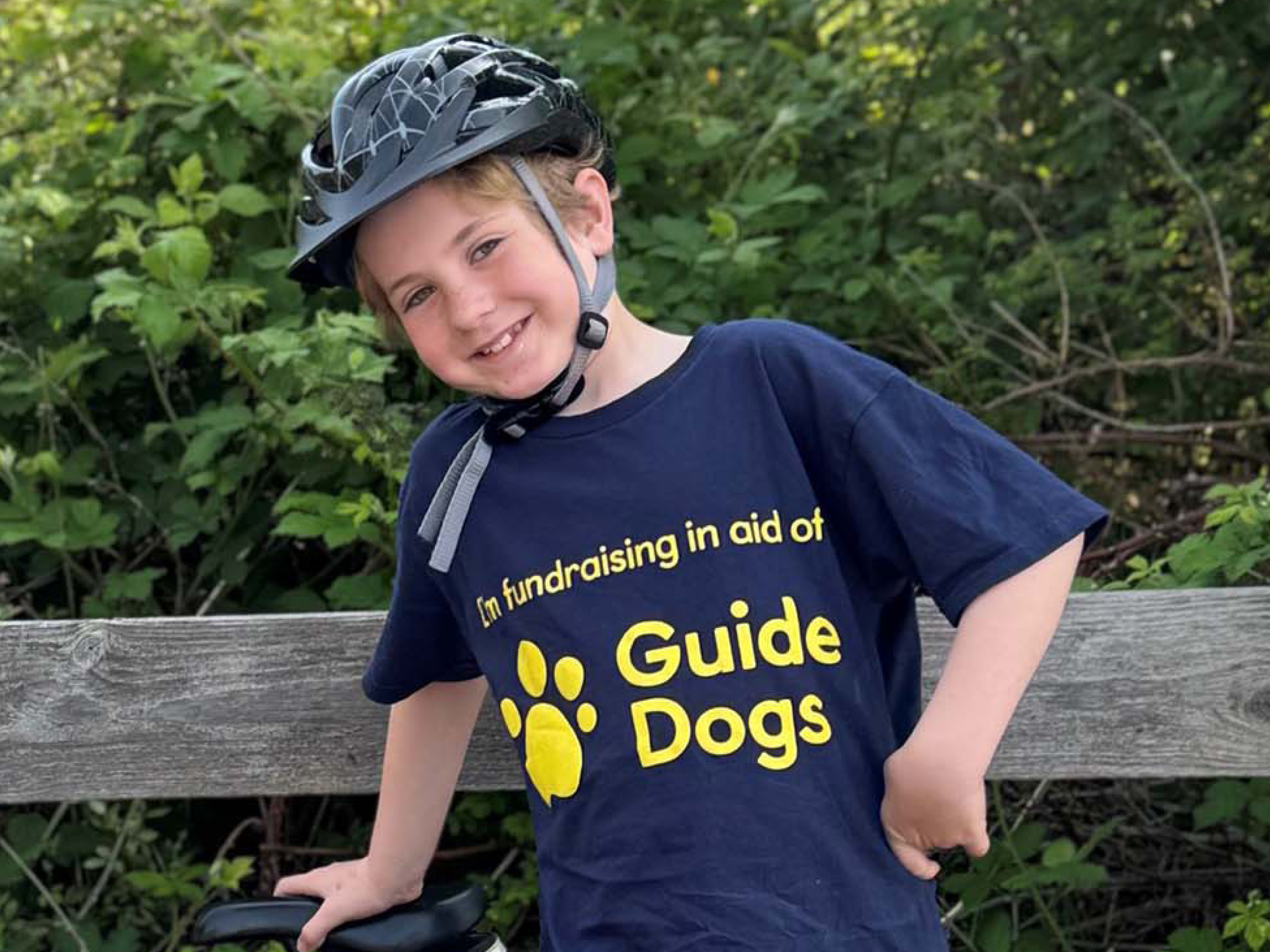 A young fundraiser called Ollie is wearing a bicycle helmet while stood up and resting his hand on the bike seat.