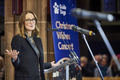 Susie Dent speaks at the front of Manchester Cathedral, with a Guide Dogs Christmas Wishes Concert banner behind her.