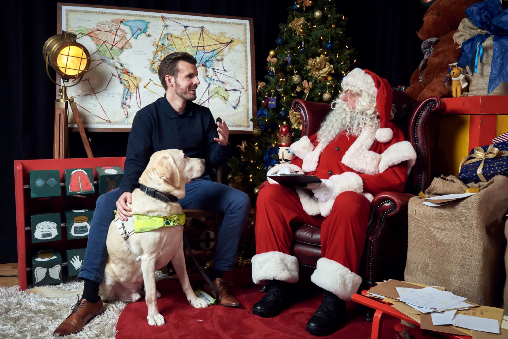 Alex Pepper, Head of Accessibility at Guide Dogs, sits with guide dog River and Santa in a Christmas grotto 