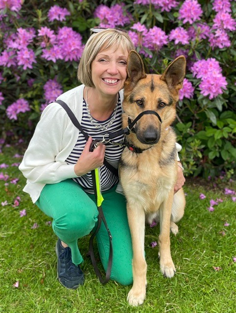Elaine smiles and crouches next to her tan German shepherd guide dog Arthur, in front of a purple rhododendron.