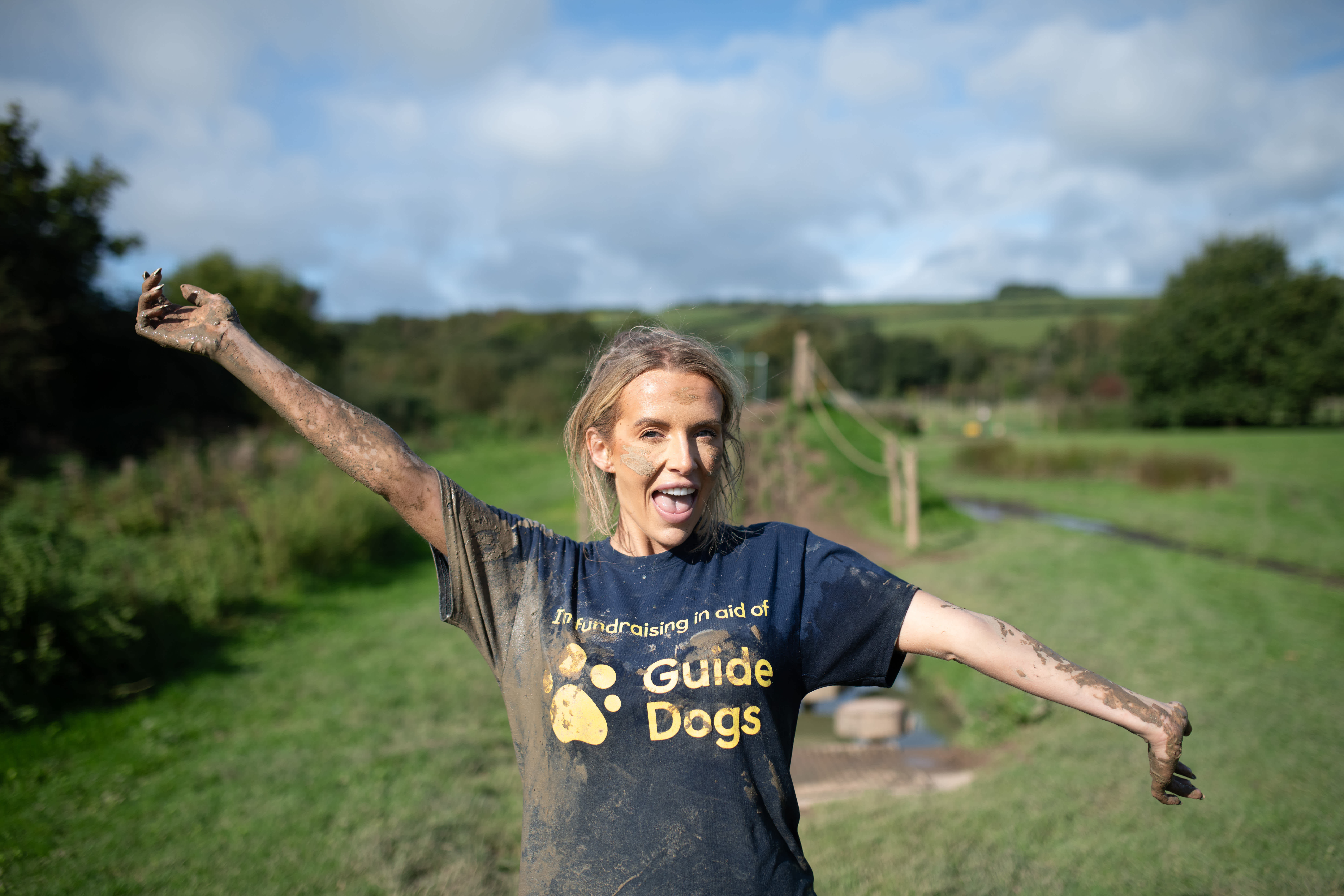 Faye Winter stands in front of an outdoor assault course. She is covered in mud and wearing her Guide Dogs t-shirt as she smiles towards the camera and holds her arms out. 