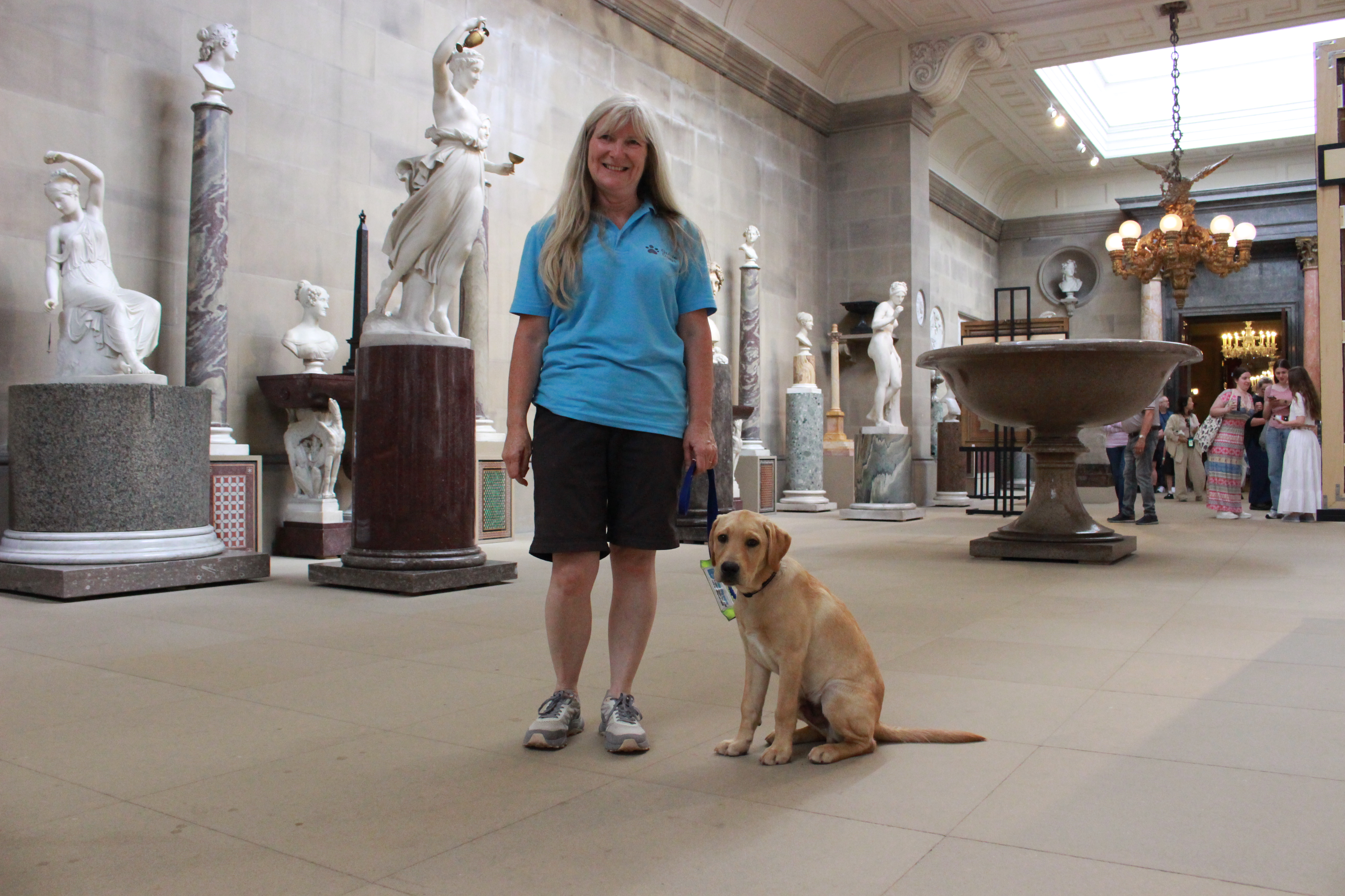 A volunteer puppy raiser and guide dog puppy stand in a room in Chatsworth House, amongst lots of marble statues and sculptures. 