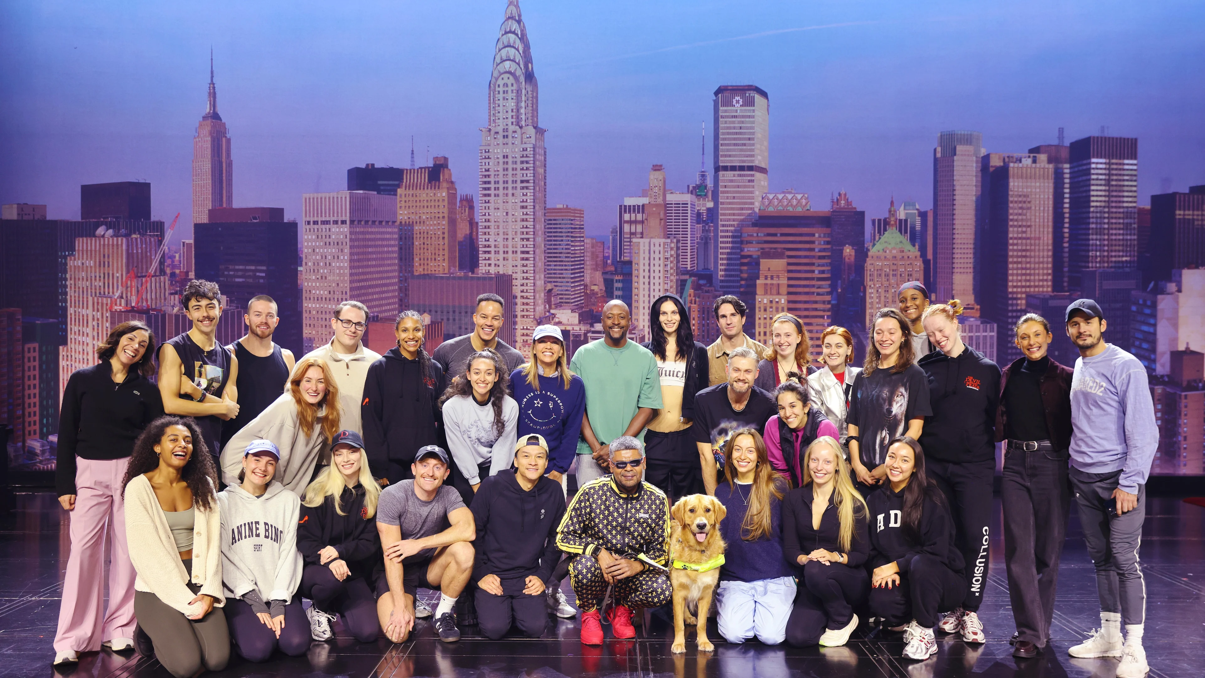 The cast of the Devil Wears Prada West End Show gathers on stage with guide dog Bill and guide dog owner Mo. They stand in front of a New York City backdrop.