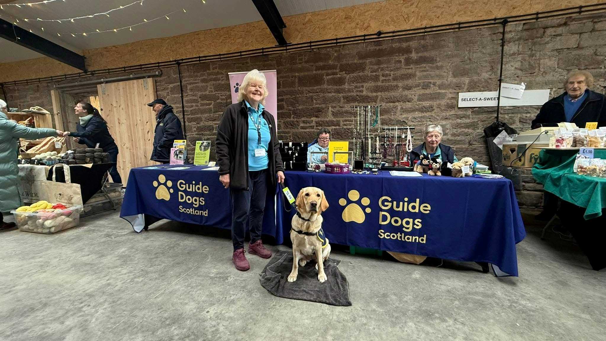 Puppy raisers Elaine stands with her yellow Labrador guide dog Lottie in front of a Guide Dogs fundraising table.