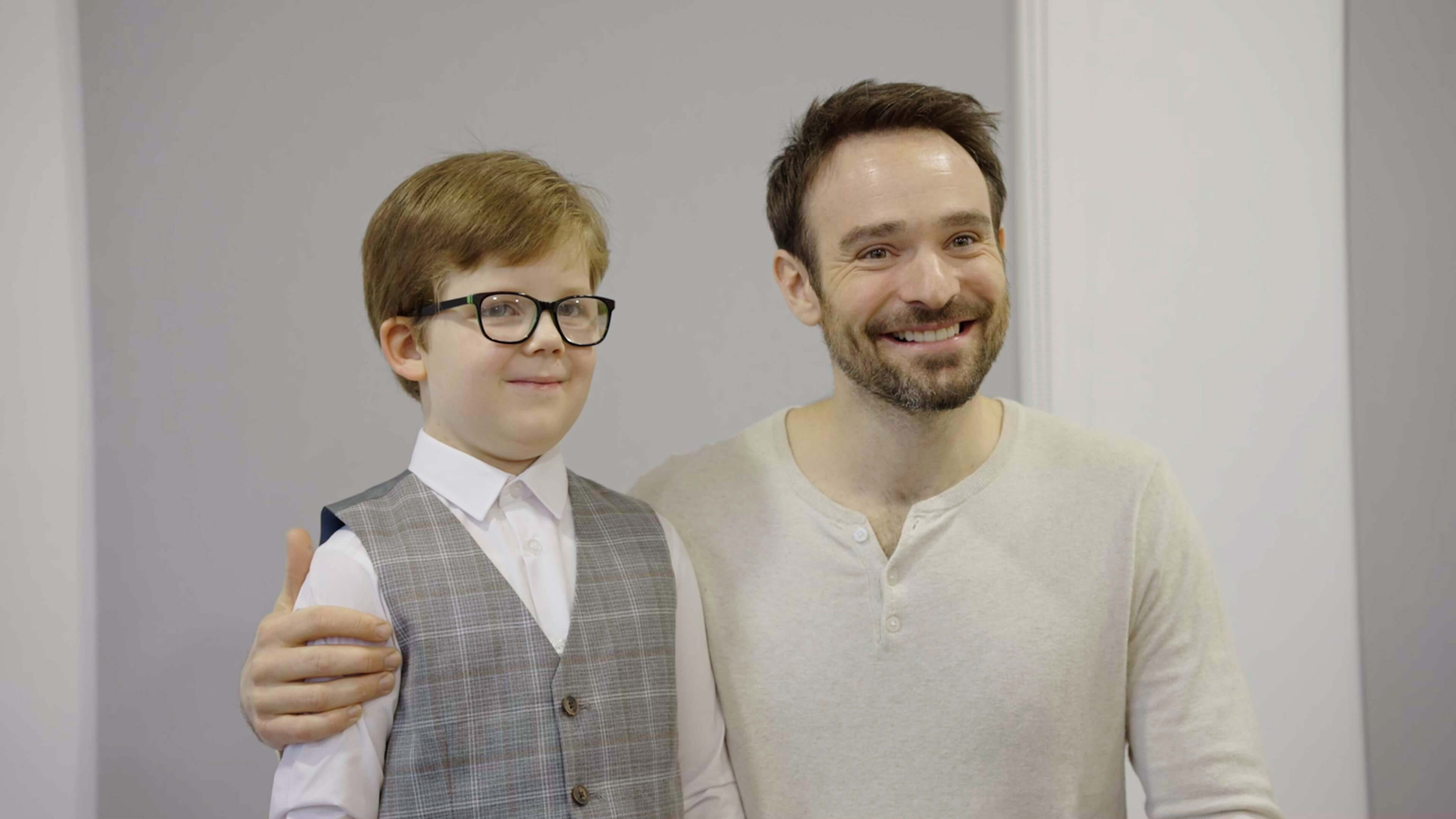 Six-year-old Rex Davies, wearing glasses, a white shirt and a grey waistcoat, stands next to Charlie Cox, who is knelling next to him. They smile to the camera.