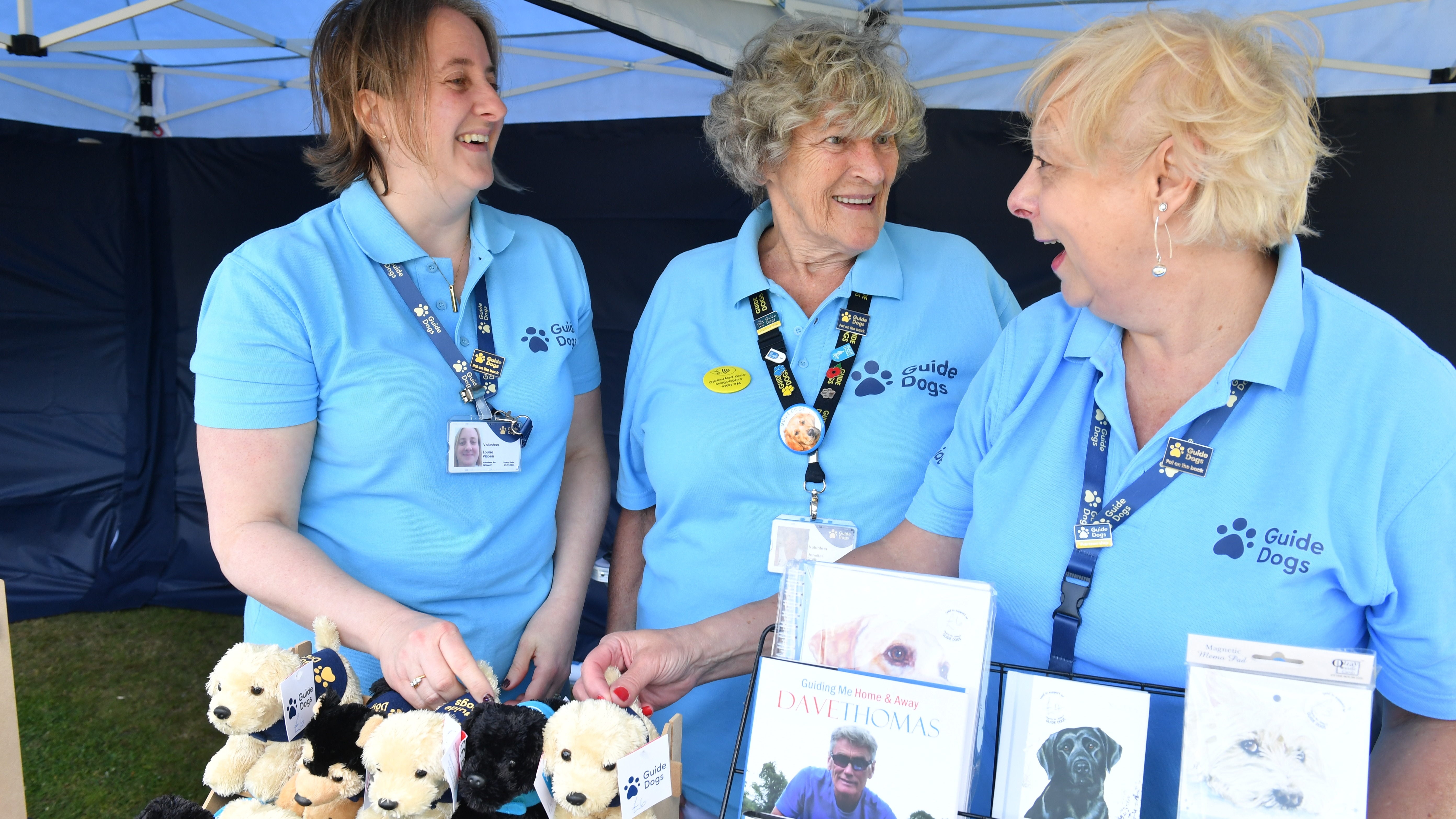 Three women in light blue Guide Dogs t-shirts smile and laugh. They are standing in a large marquee behind a table with Guide Dogs soft toys and a selection of books about Guide Dogs.