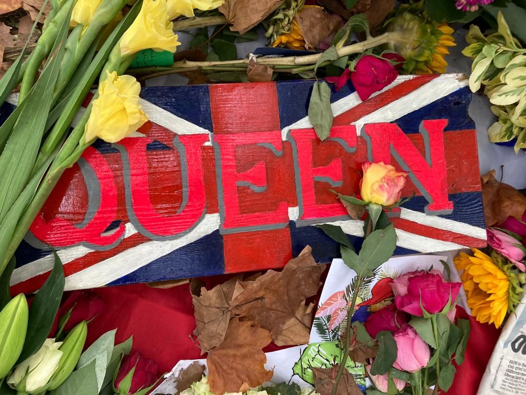A close-up of some of the flowers and notes left in Green Park, London, in memory of HM The Queen. The display includes a Union flag with word 'Queen' written in large, red letters across it. 