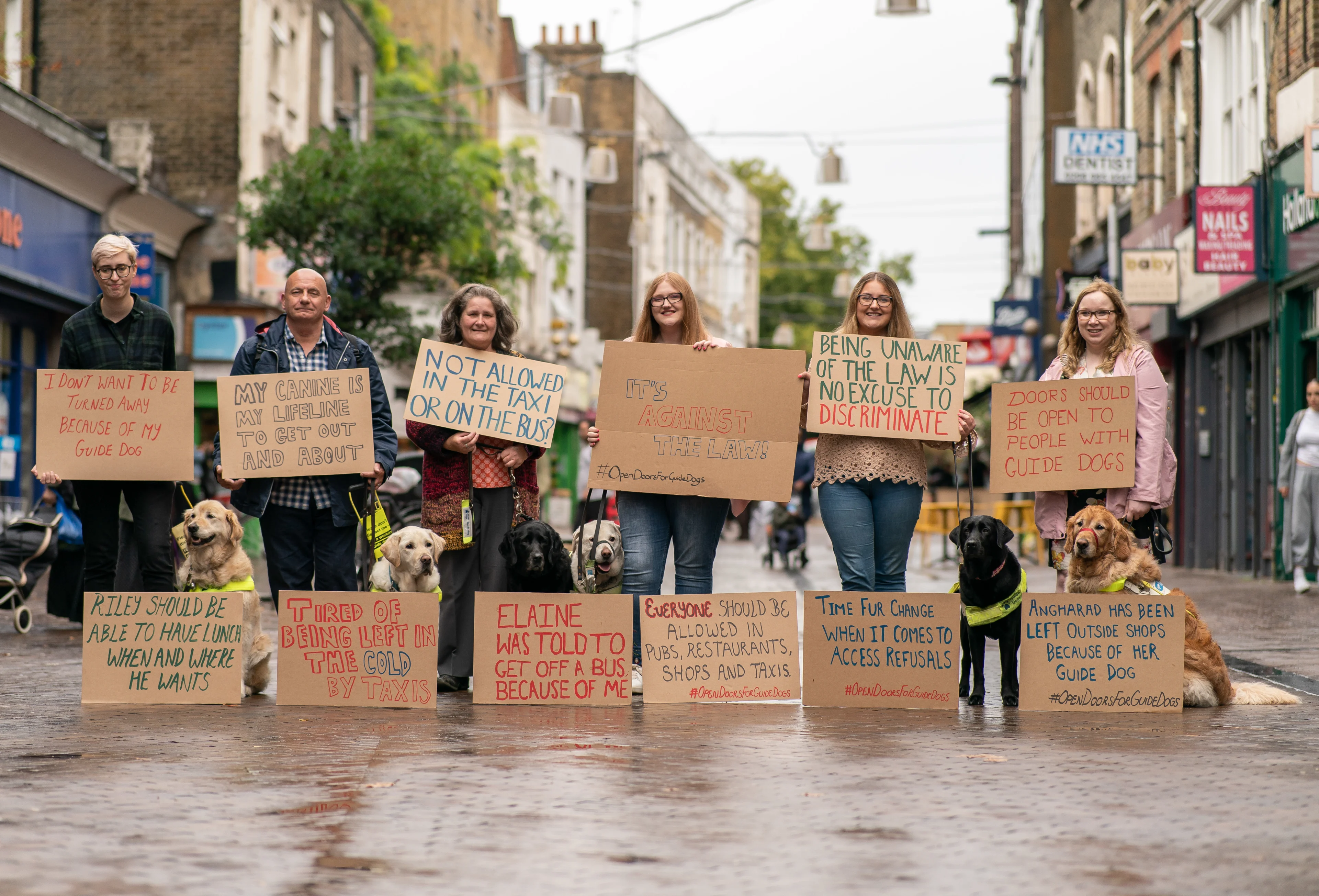 Guide dog owners and their guide dogs gather with signs for Open Doors 