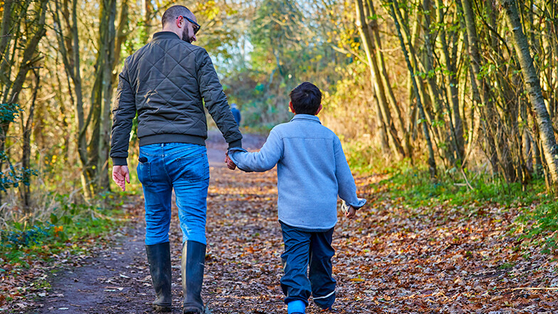 Dad Phil walking and holding hands with his young son Teddy on a sunny woodland pathway.