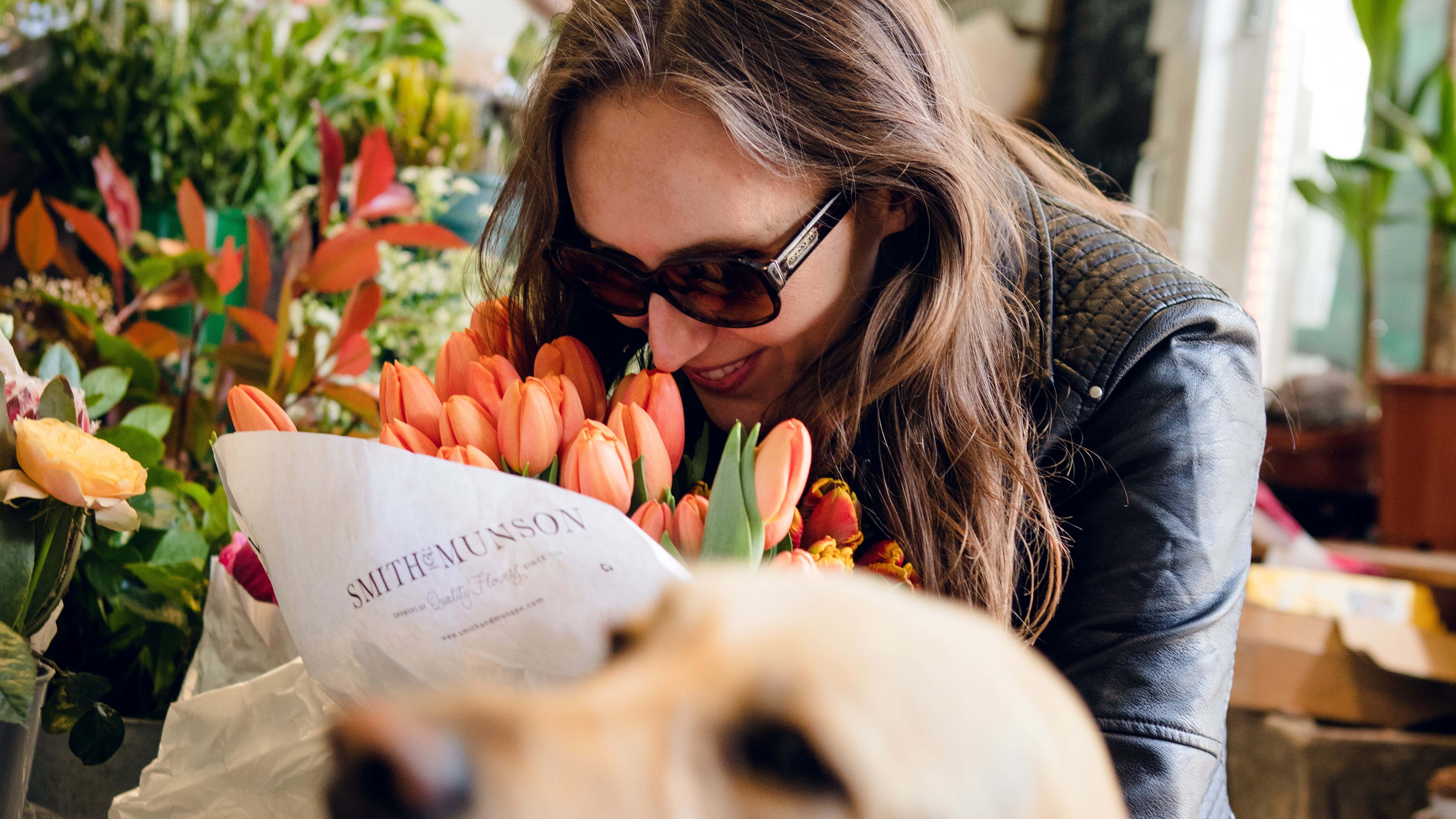 Female guide dog owner with long dark hair and sunglasses sniffs a bouquet of orange tulips, while standing in a flower shop. A yellow Labrador's head is in the foreground, out of focus.