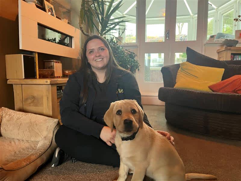 Puppy Development Advisor Terri sits on the floor of a living room with yellow Labrador puppy Ritzy. 