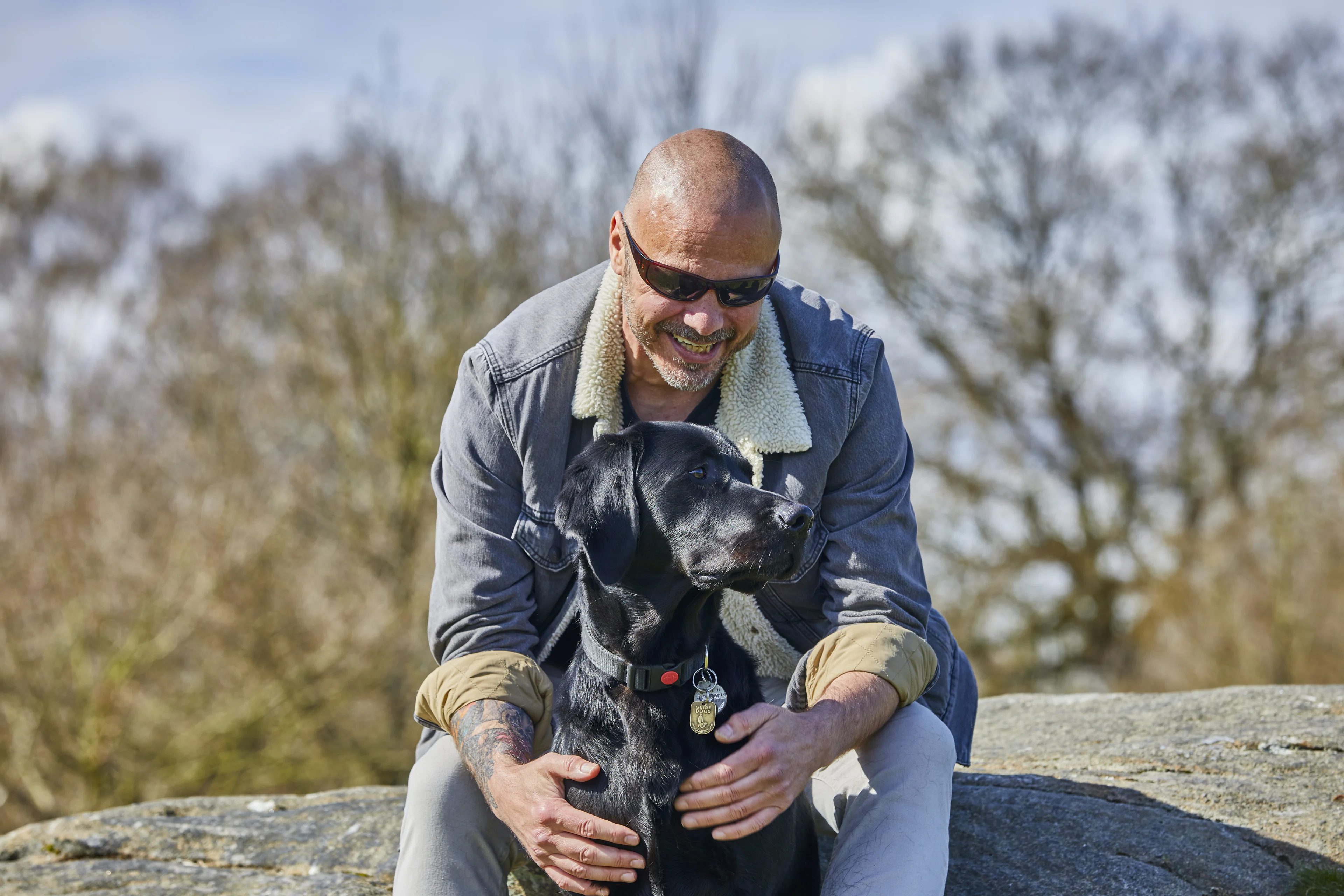 Terry sits on a rock and smiles down at black Labrador cross guide dog Spencer, who is sitting between Terry's knees