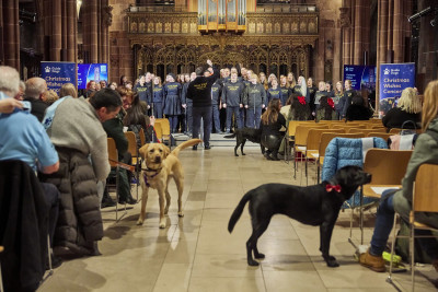 A view down the aisle of Manchester Cathedral. A choir sings at the front while the audience watches and dogs stand in the aisle.