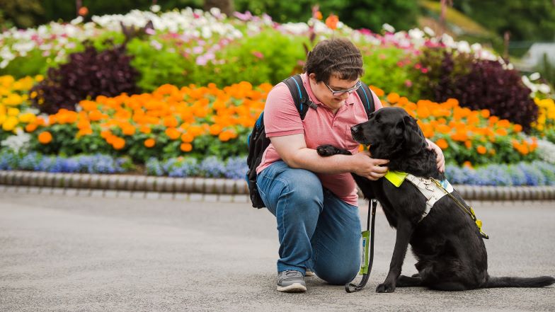 A guide dog owner kneels down next to their black guide dog in front of a flower bed  