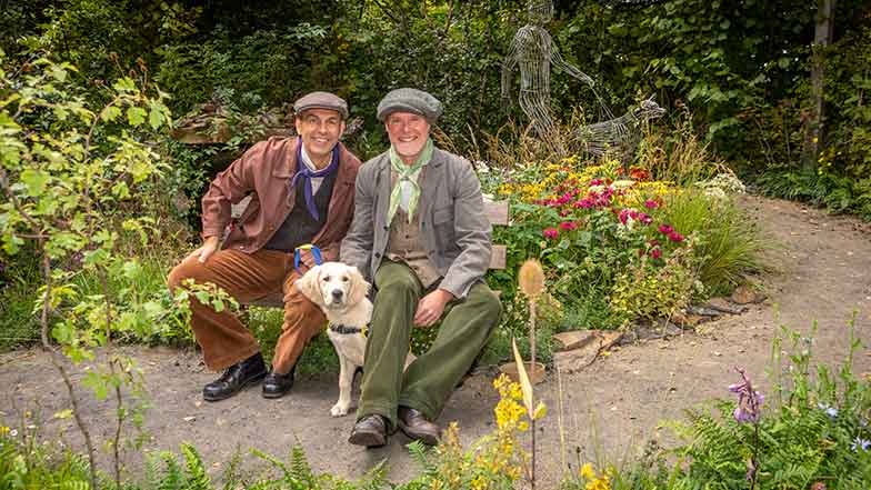 Adam-Woolcott-and-Jonathan-Smith, in period costume, sitting in the Gude Dogs award winning Chelsea Flower Show Garden with a trainee guide dog