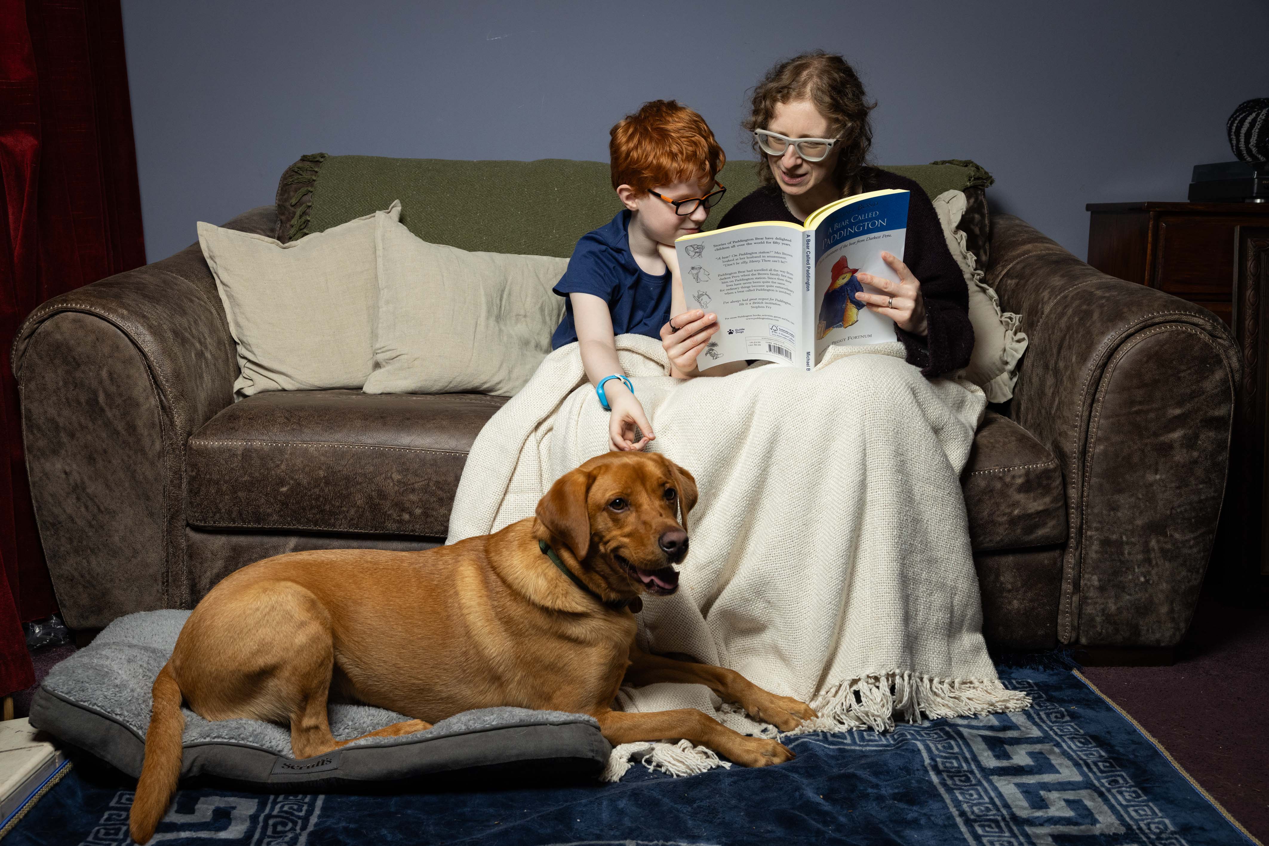 A mum and her son sit side by side on the sofa reading 'Paddington Bear'. The boy is leaning down slightly to stroke the top of his dog's head who is lying on a dog bed by their feet. 