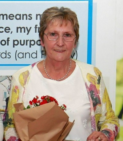 Sue, who has short blonde hair and glasses, wears a green and red floral cardigan and holds a bouquet of red flowers in brown paper.