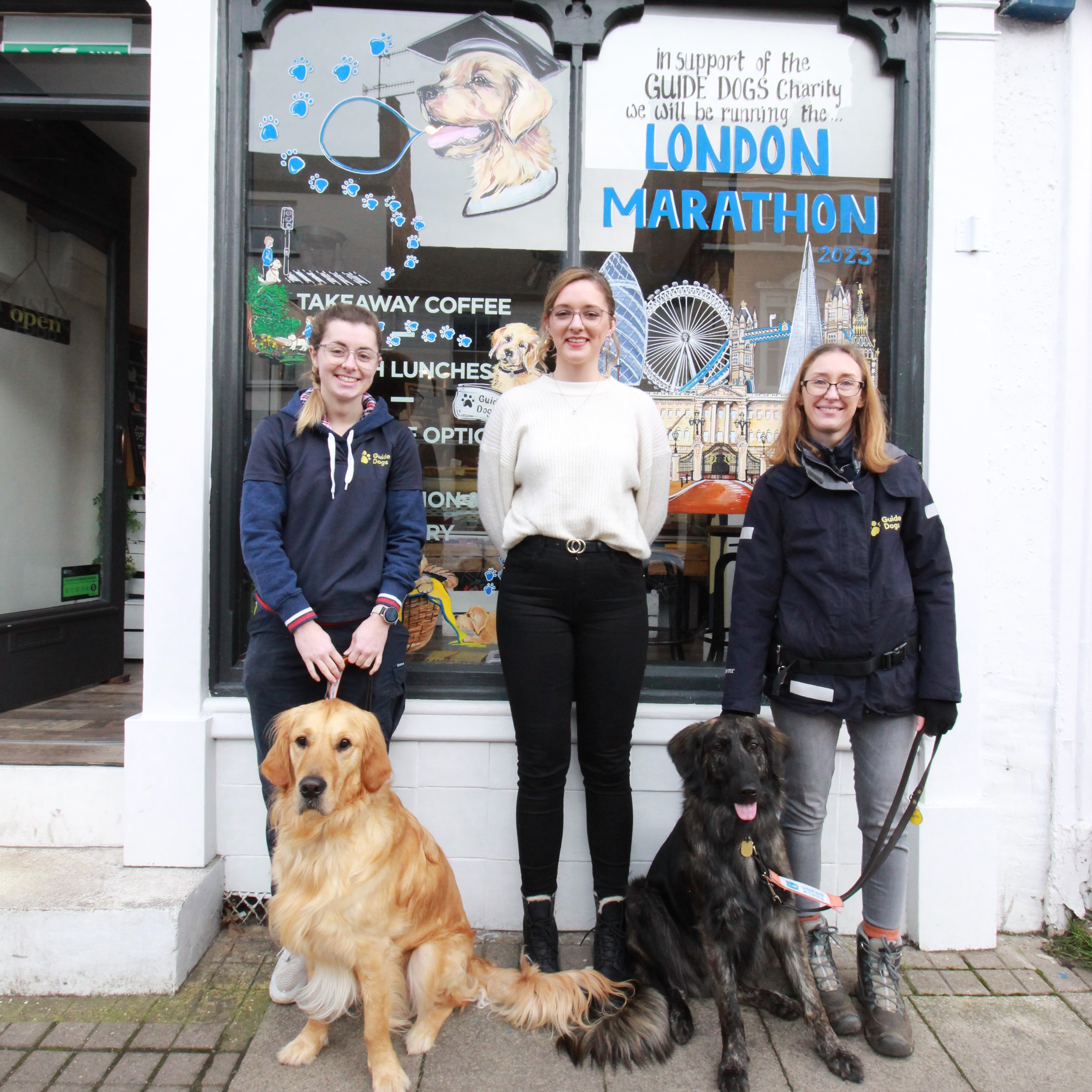 Photo shows guide dog trainer Gemma, Alice from Rustic Food and guide dog trainer Katie, with Ashleigh, a Golden Retriever cross German Shepherd and Charlie, a Golden Retriever, stood outside Rustic Food in front of the window mural. The mural shows a series of dog images including puppies in a basket, and a dog wearing a graduation hat, and the London skyline below the words London Marathon. 