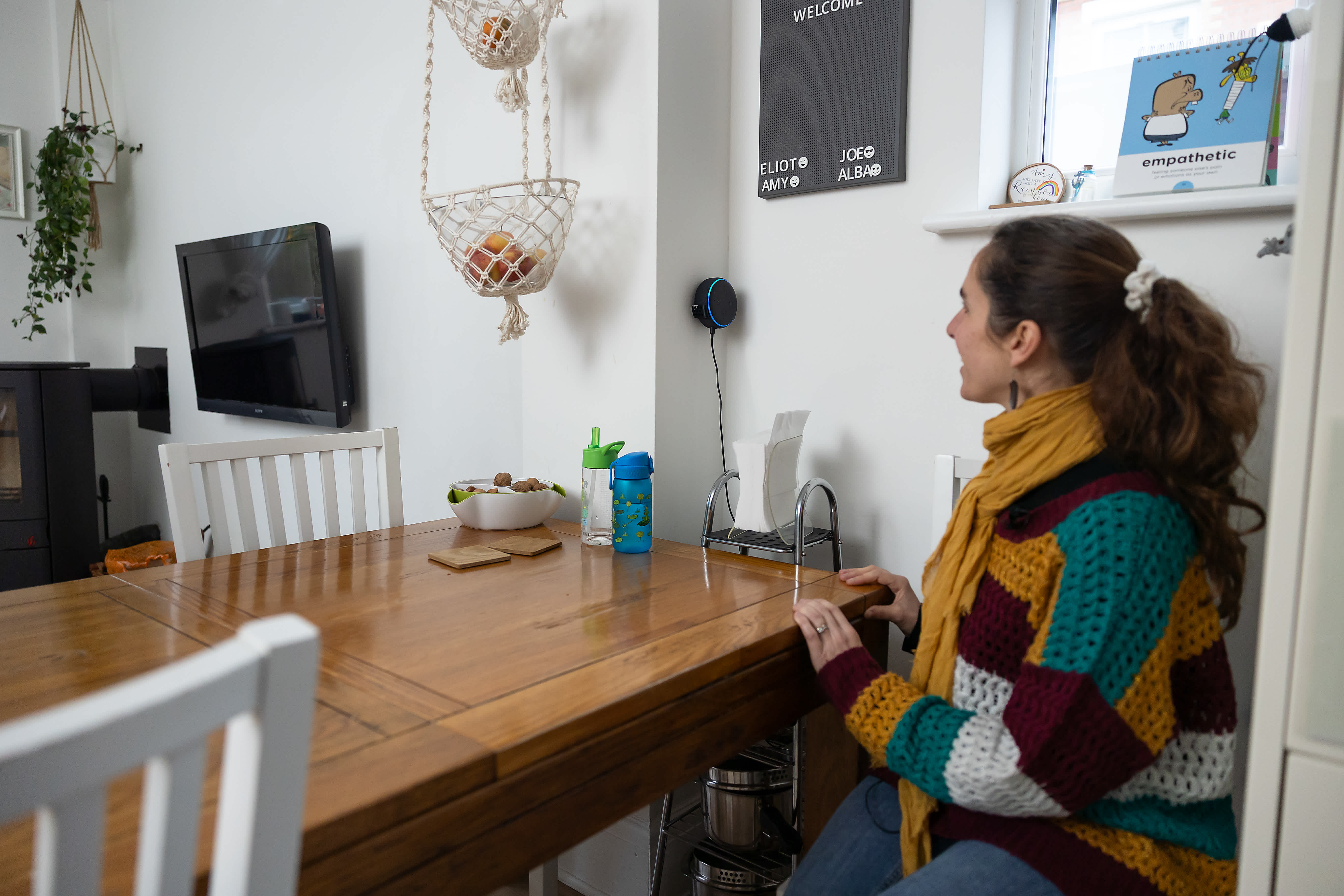 A woman is sat at a table in her kitchen, giving voice instructions to her Amazon Echo smart speaker. 