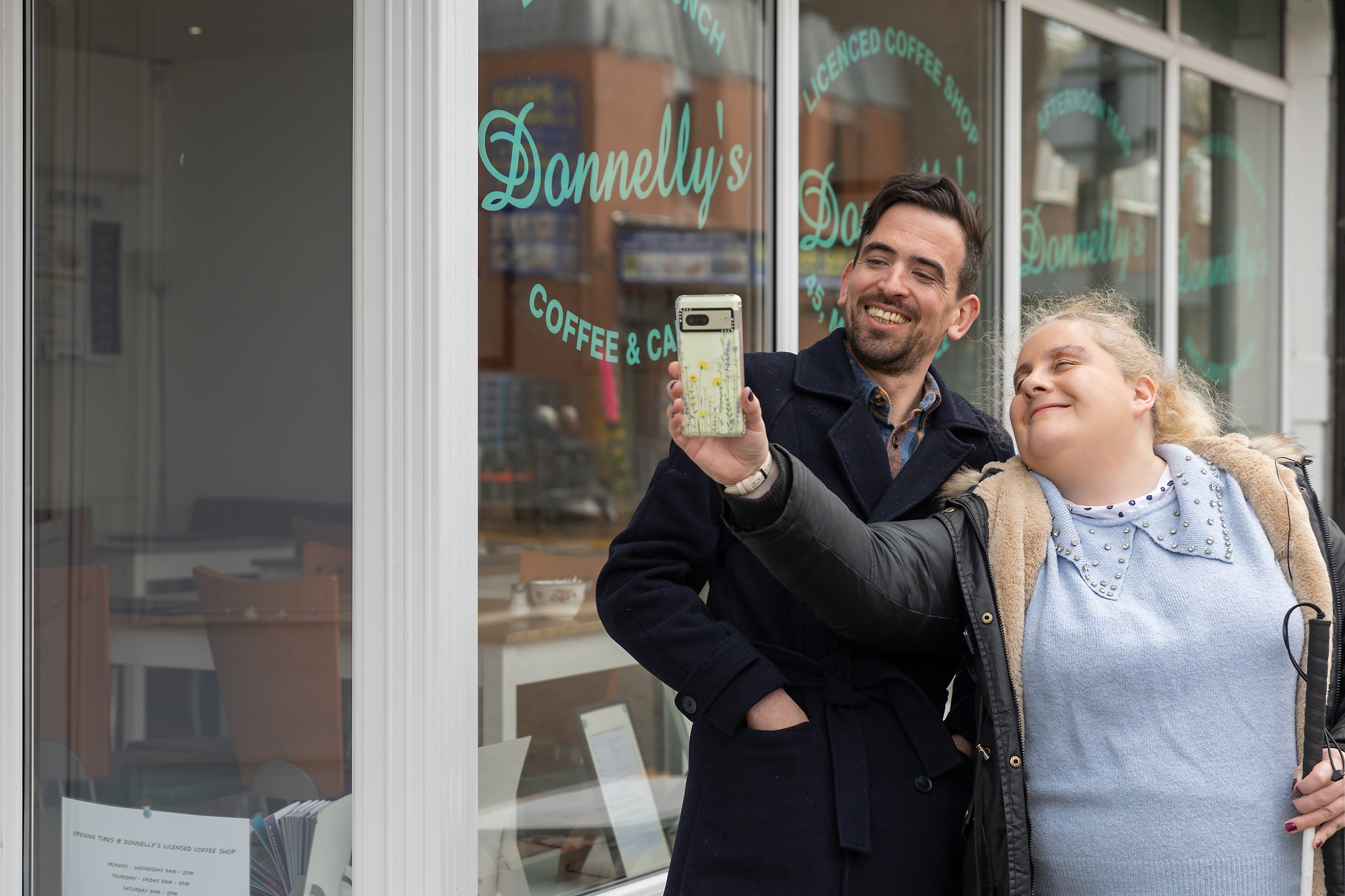 Two people use the Google Pixel phone to take a selfie outside of a cafe. They both smile into the camera as the woman holds her phone in one hand and a white cane in the other. 