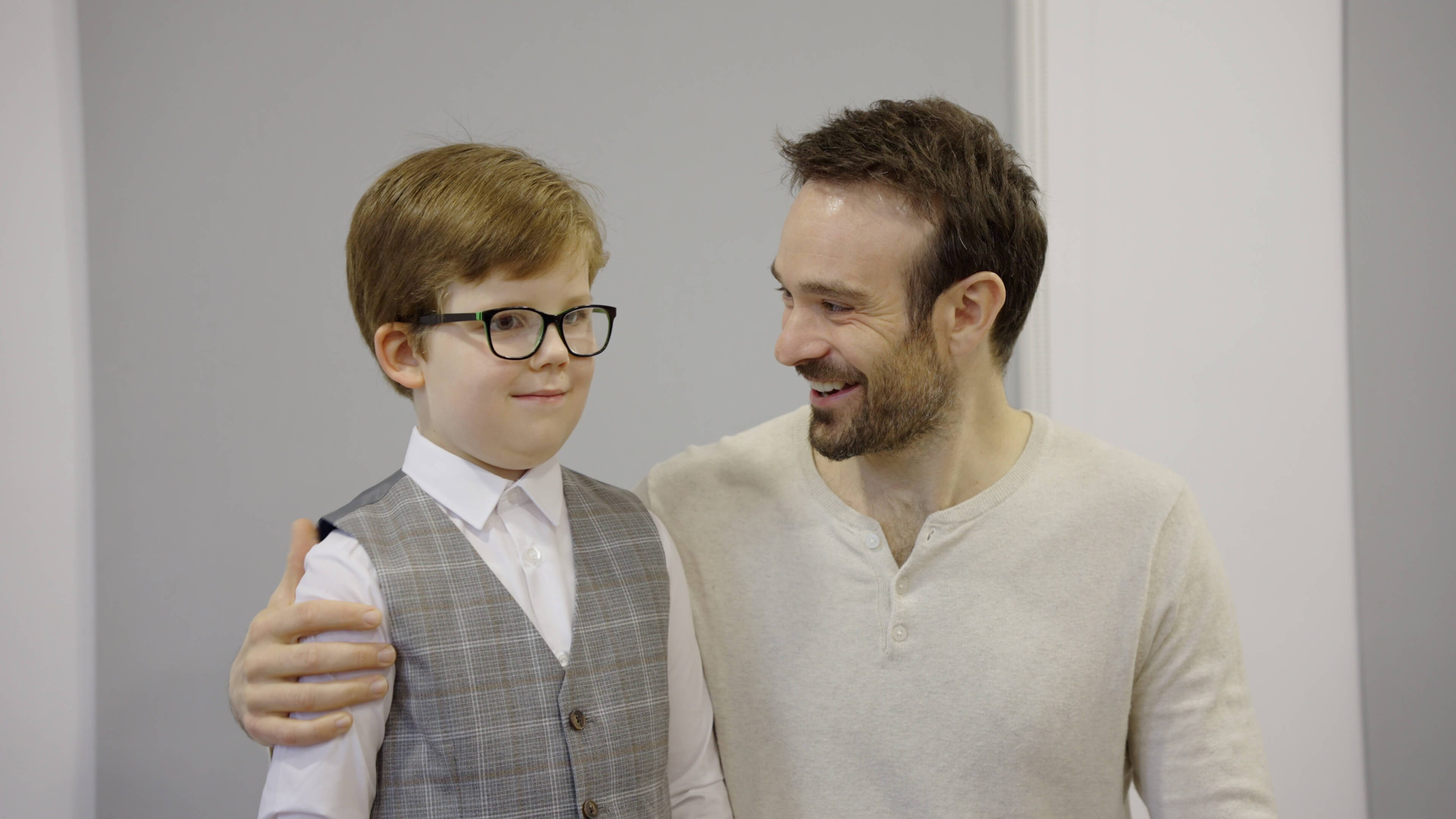 Six-year-old Rex, wearing glasses, a white shirt and grey waistcoat, stands next to Charlie Cox, who is kneeling next to him with his arm around him. Charlie looks at Rex and smiles.