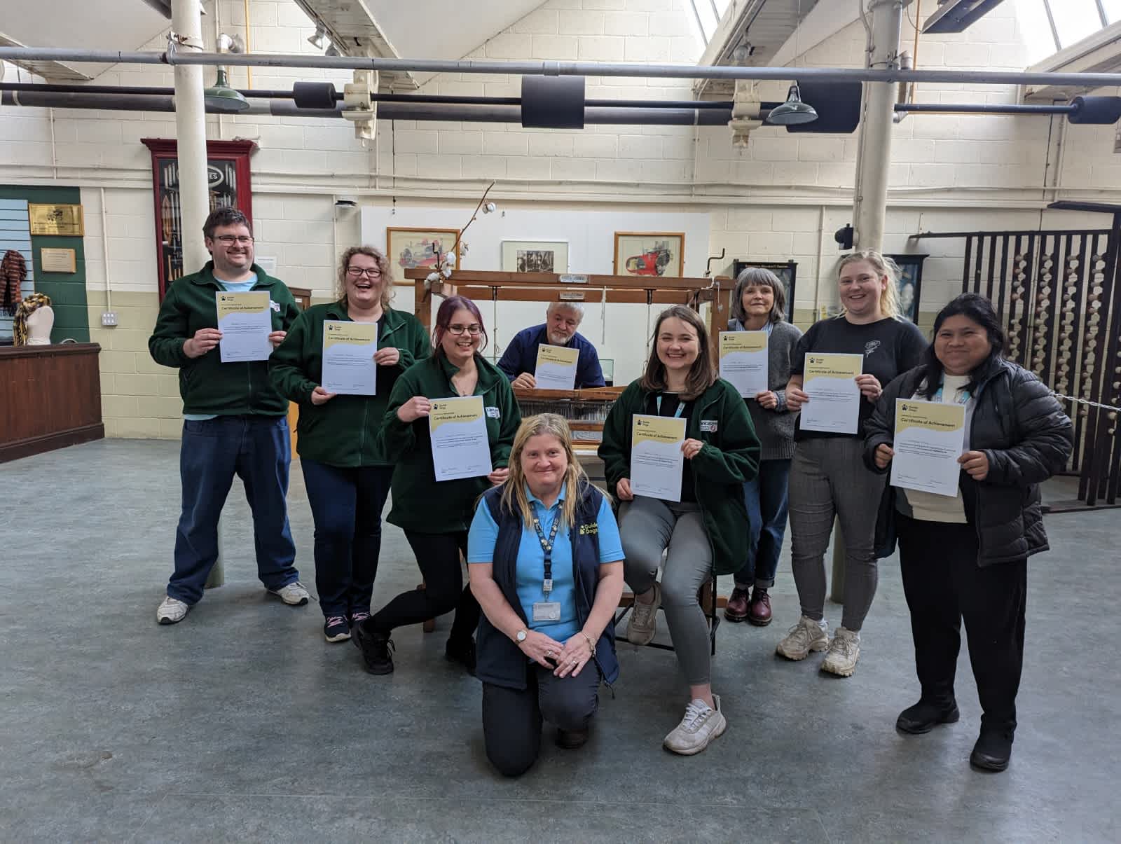Sharon from Guide Dogs is crouching in front of staff from Queen Street Textile Museum, who are holding up their certificates.