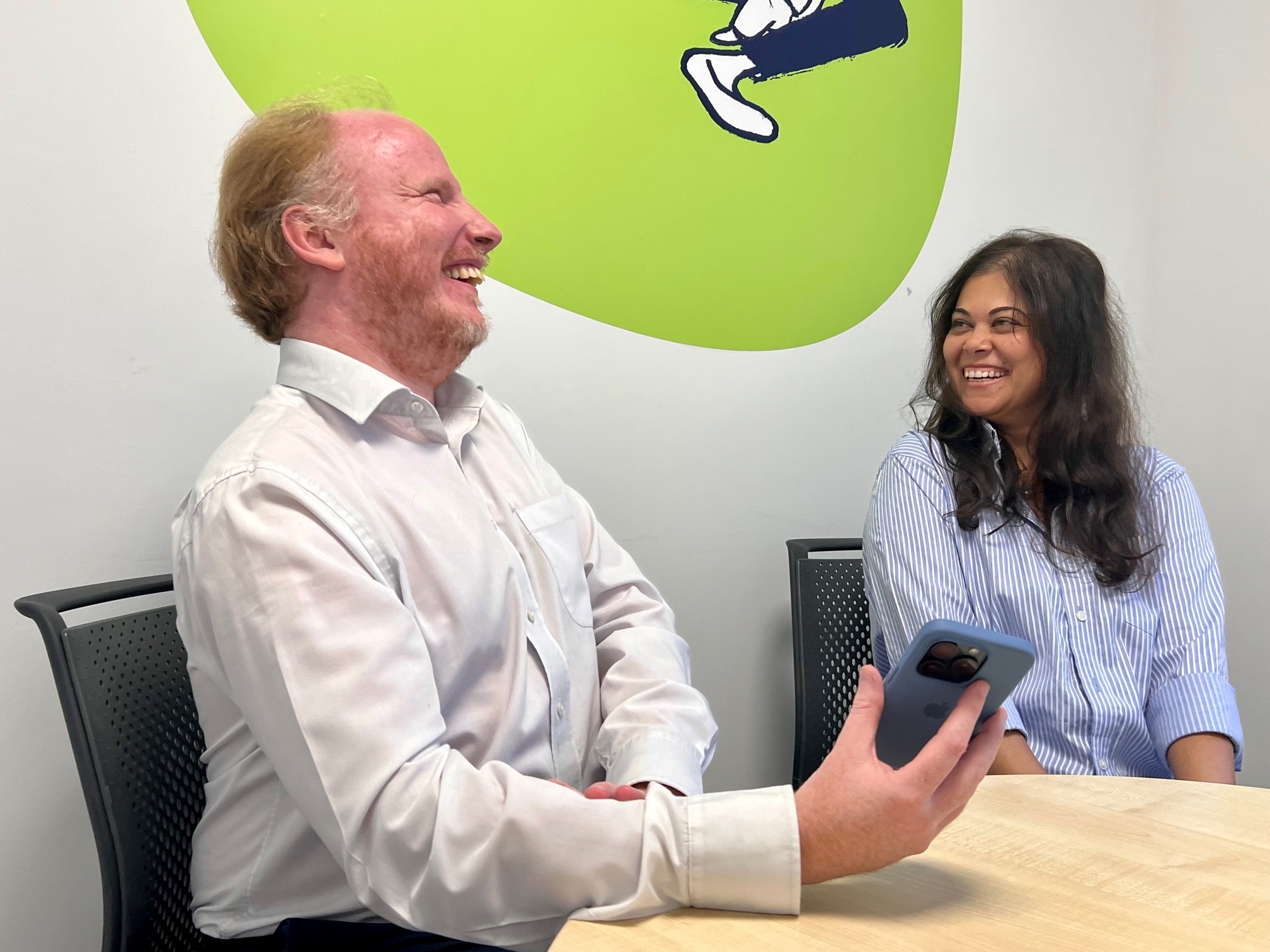 Darren and Pooja, who are sat together at a table, are laughing as they sharing a joke. Darren, who has sight loss, is holding an iPhone.