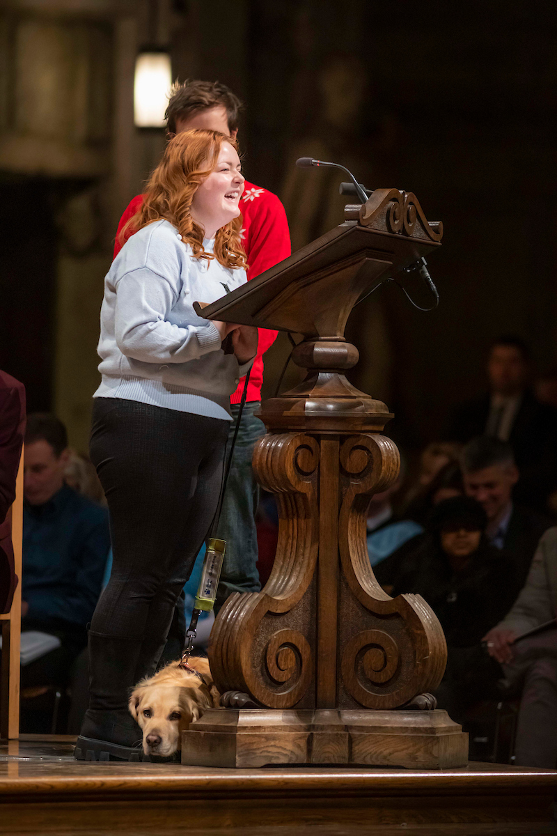 Lucy Edwards telling her sight loss story at the lectern in St Paul's Cathedral, with guide dog Molly at her feet. 