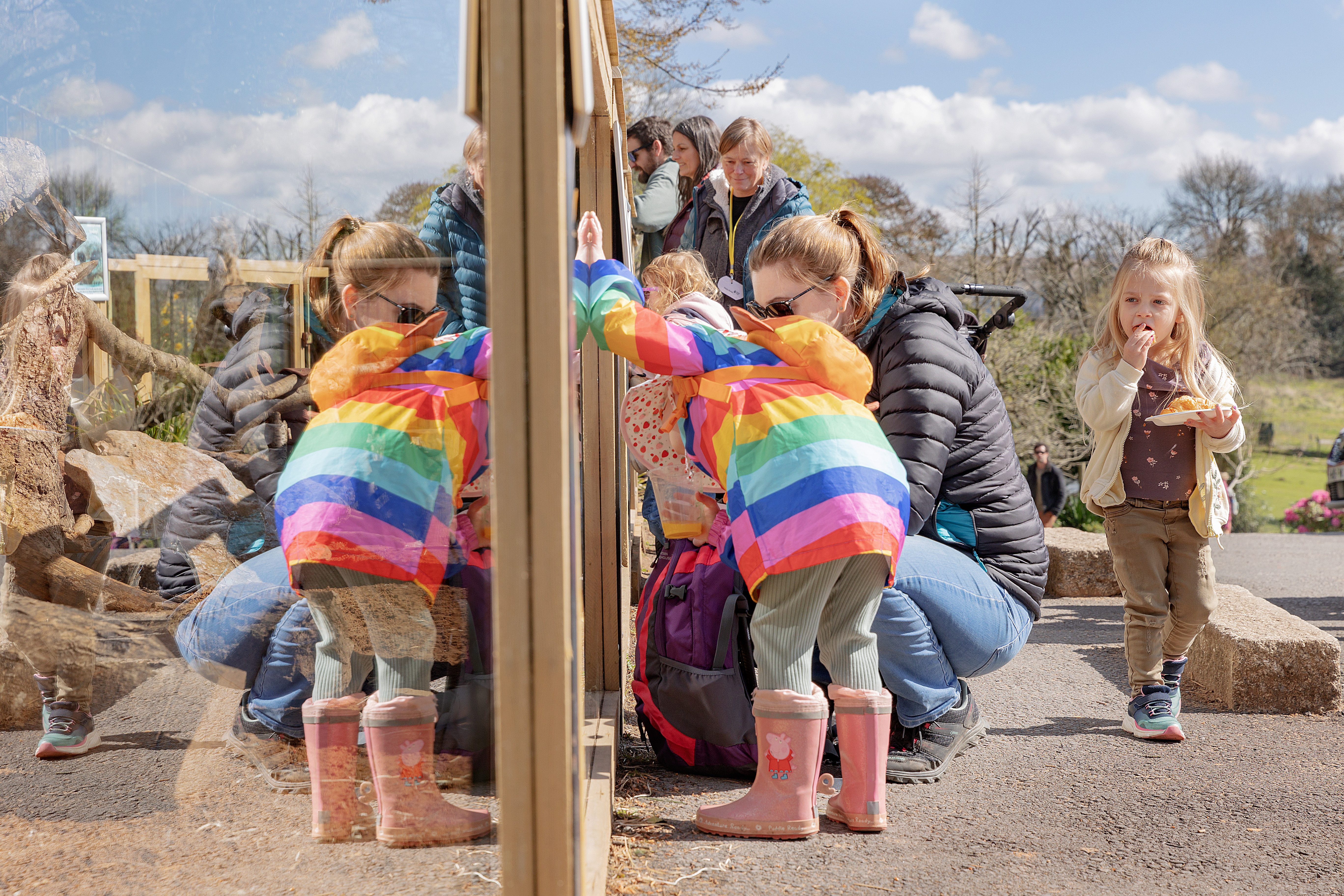 Image of children and parents at a zoo family event. The image shows a child wearing a multicoloured coat, which reflects in the glass animal enclosure.