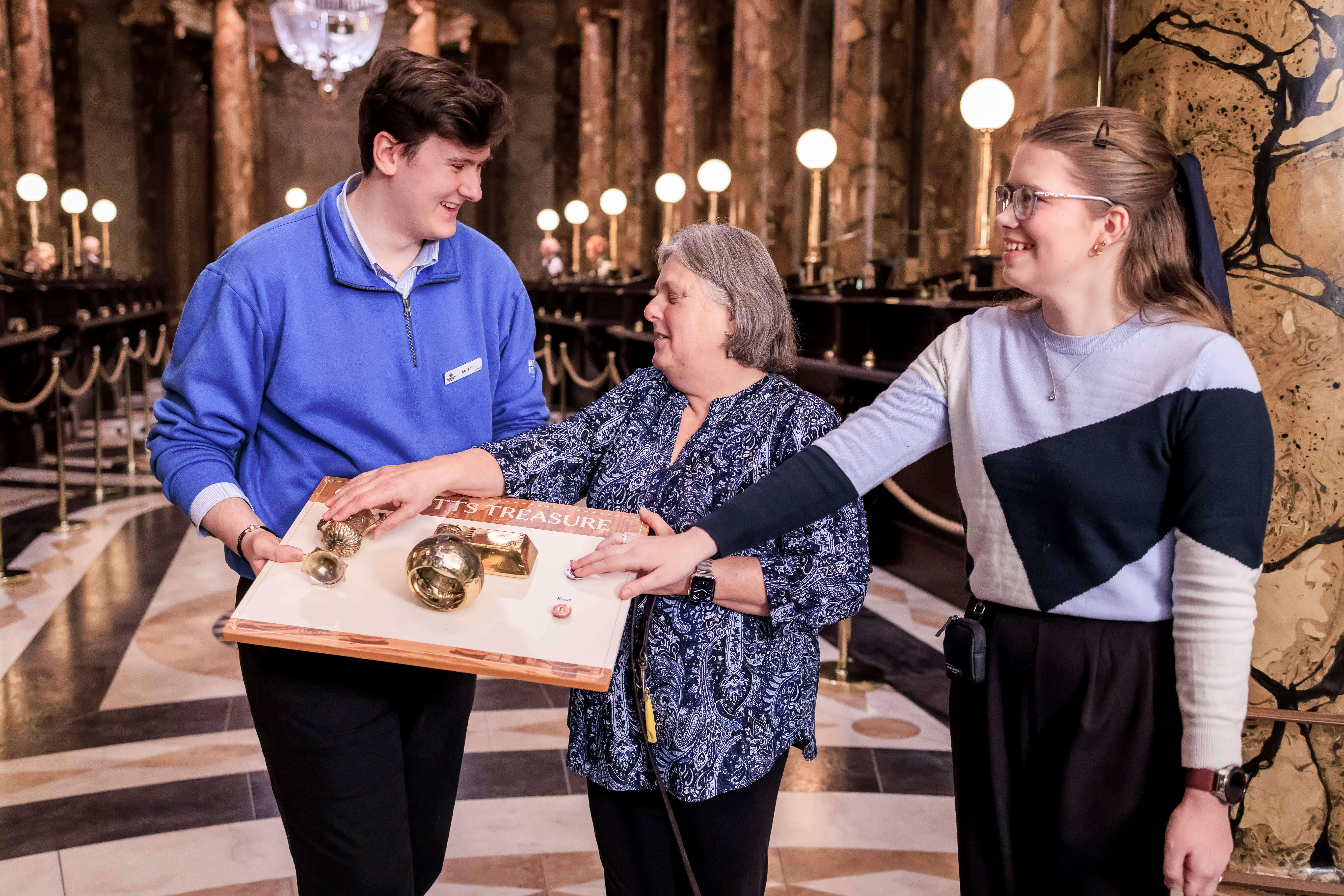 Guide dog owners Penny and Skie touch gold treasure props at Gringotts Bank inside the Warner Bros Studio Tour. The props are held on a tray by a staff member.