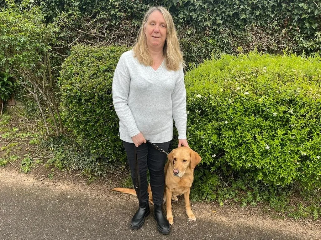 Guide dog owner Wend stands next to guide dog Clover, pictured outside in front of a hedgerow. 