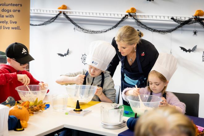 HRH The Duchess of Edinburgh meets young service users Theo, Oliver and Chloe who are making dog treats in bowls with spoons. Halloween decorations are in the background. 