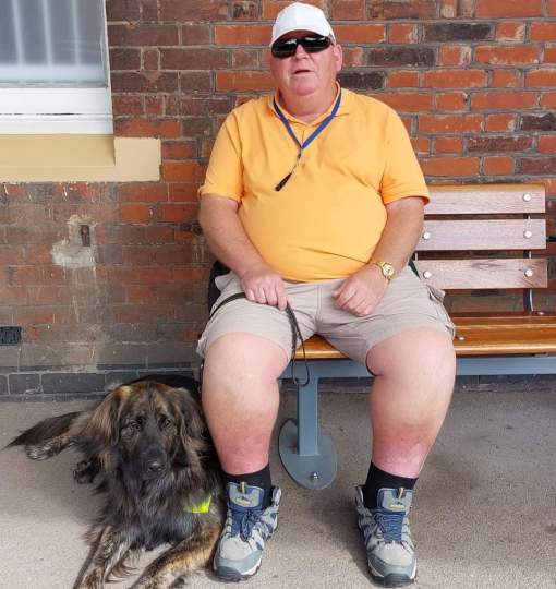 Guide dog owner Ron sat on a bench looking towards the camera. Hugo is lying on the floor next to him, wearing his white harness.