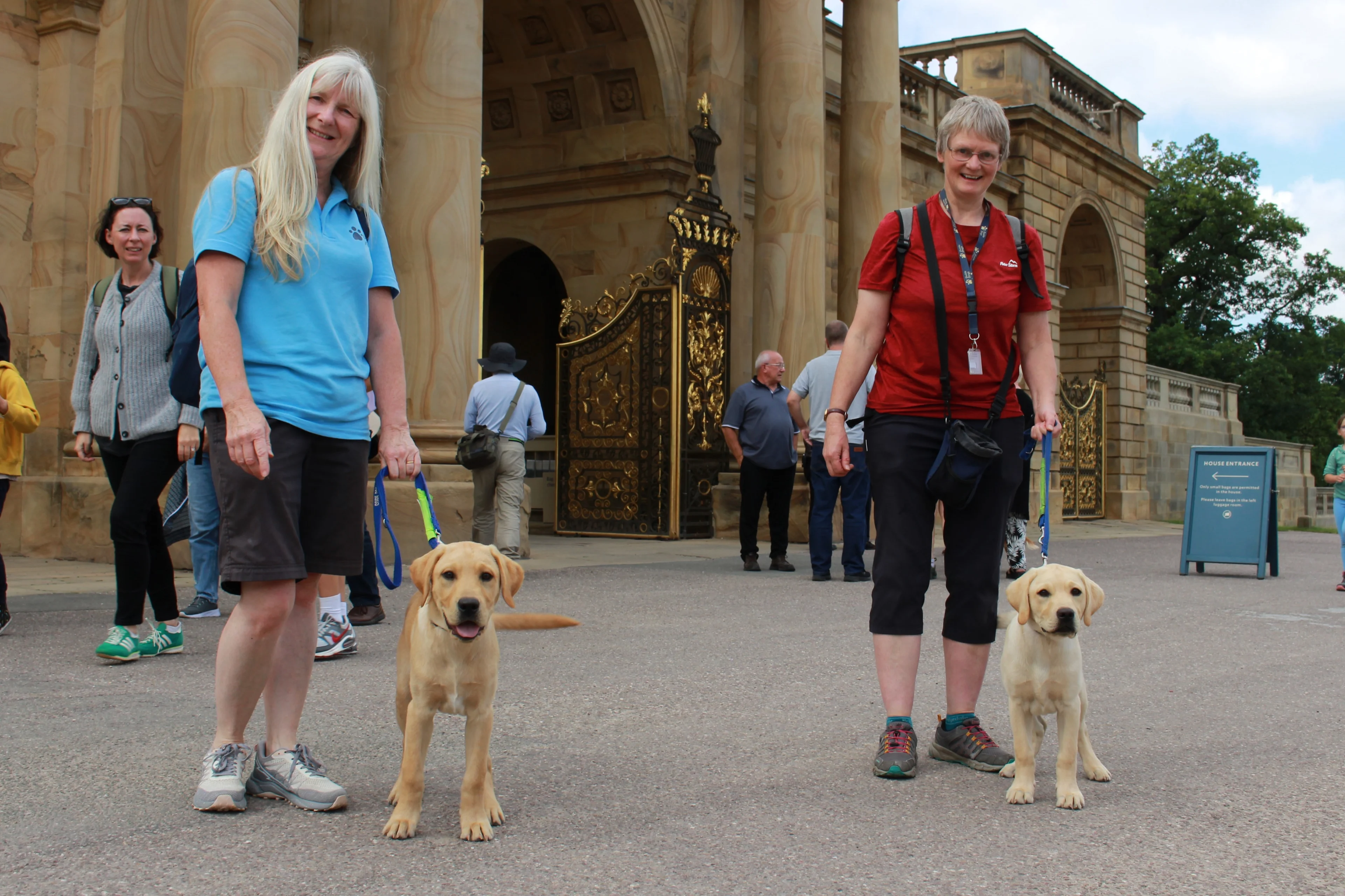 Two volunteer puppy raisers stand outside the magnificent arched entrance to Chatsworth House, as they hold the leads of two yellow Labrador puppies in training.