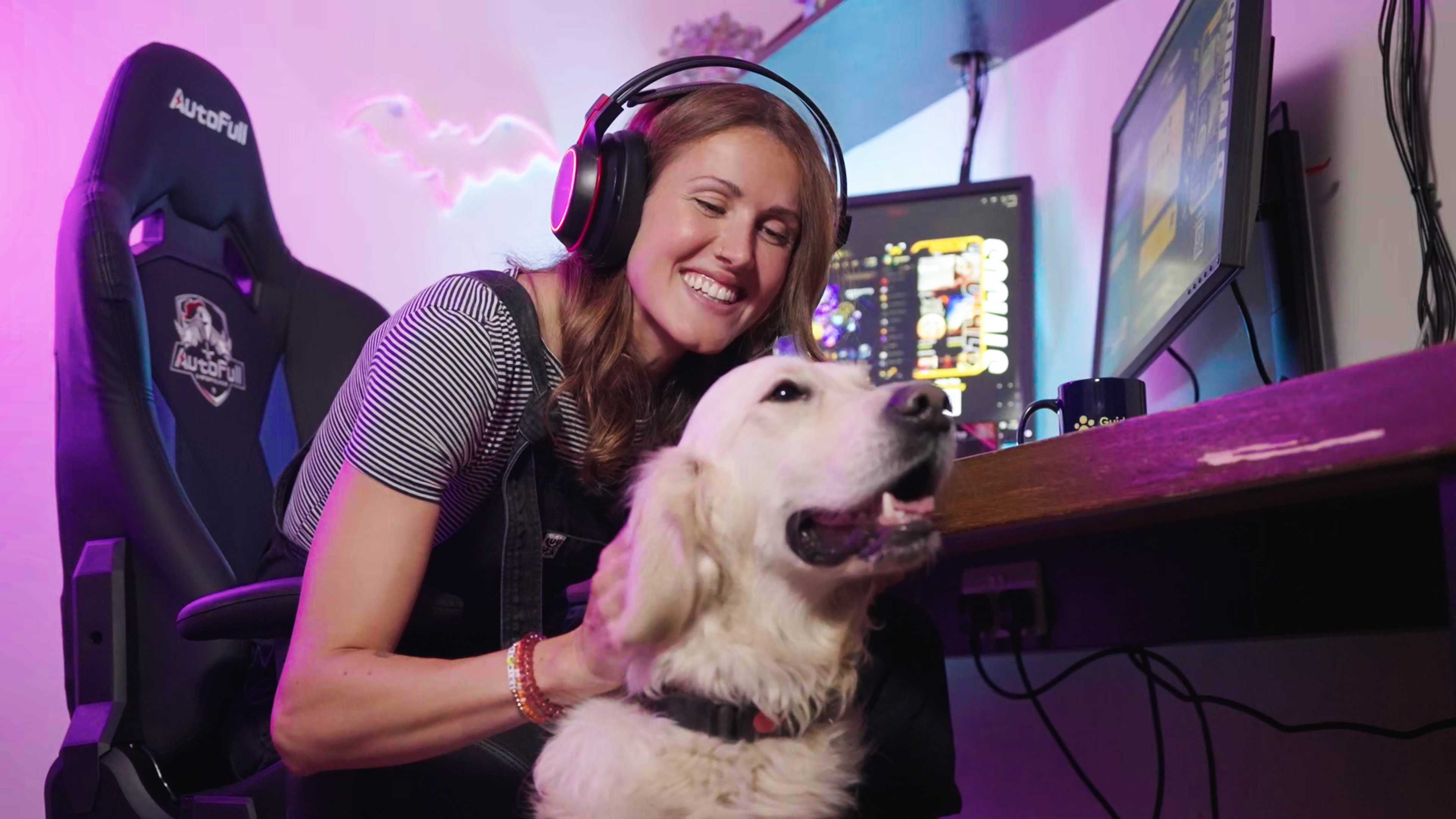 A person wearing headphones sat beside a gaming set-up and cuddling a yellow dog.