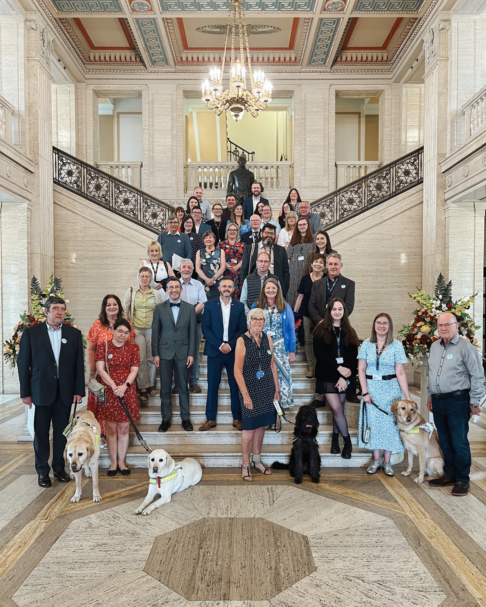 A group of staff, volunteers, service users and dogs stand on a staircase in a beautiful hall underneath a chandelier.