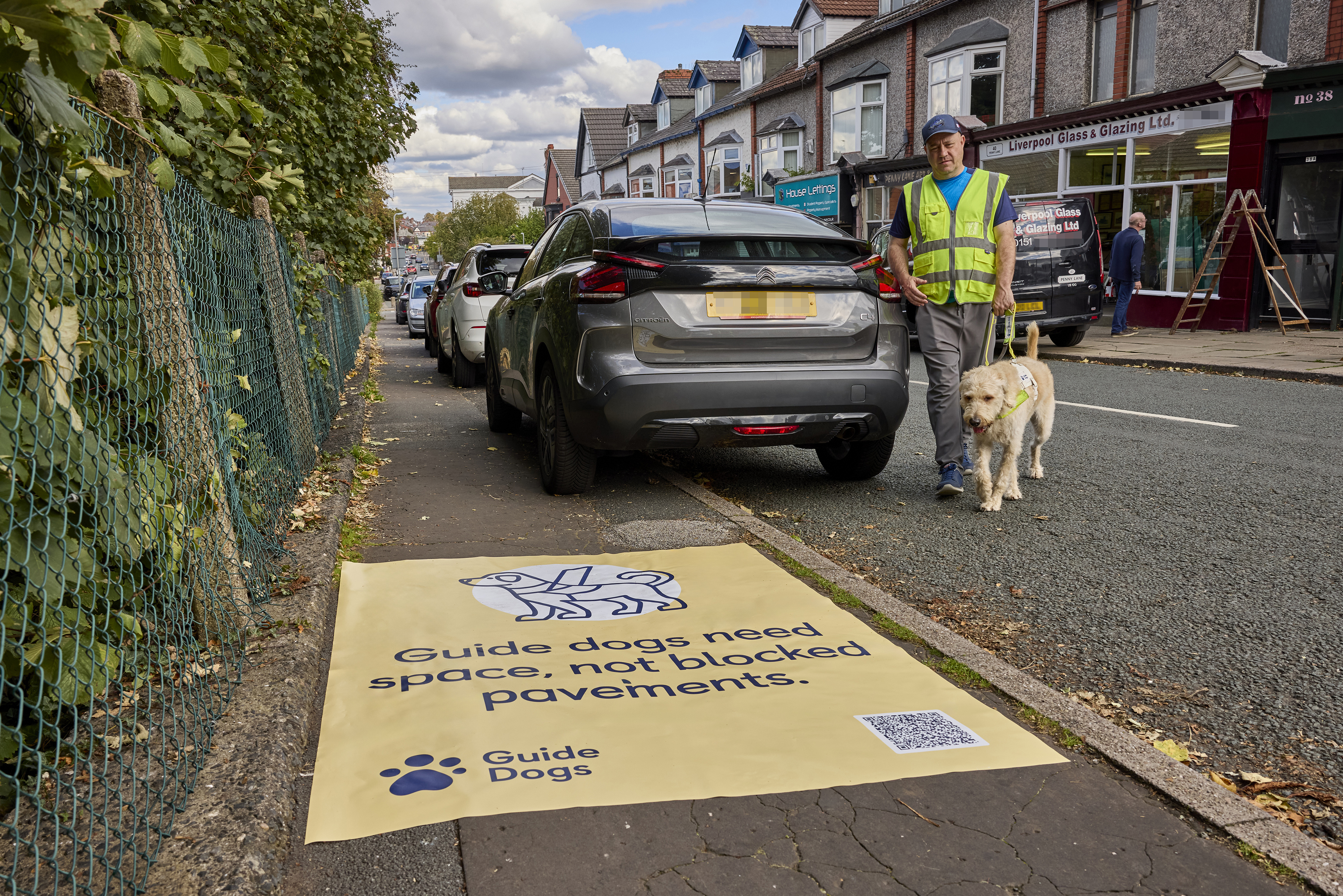 A man and his guide dog walk around the back of a car after being forced into the road because the car is parked on the pavement. In the foreground there is a yellow sticker on the pavement that says "Guide dogs need space, not blocked pavements".