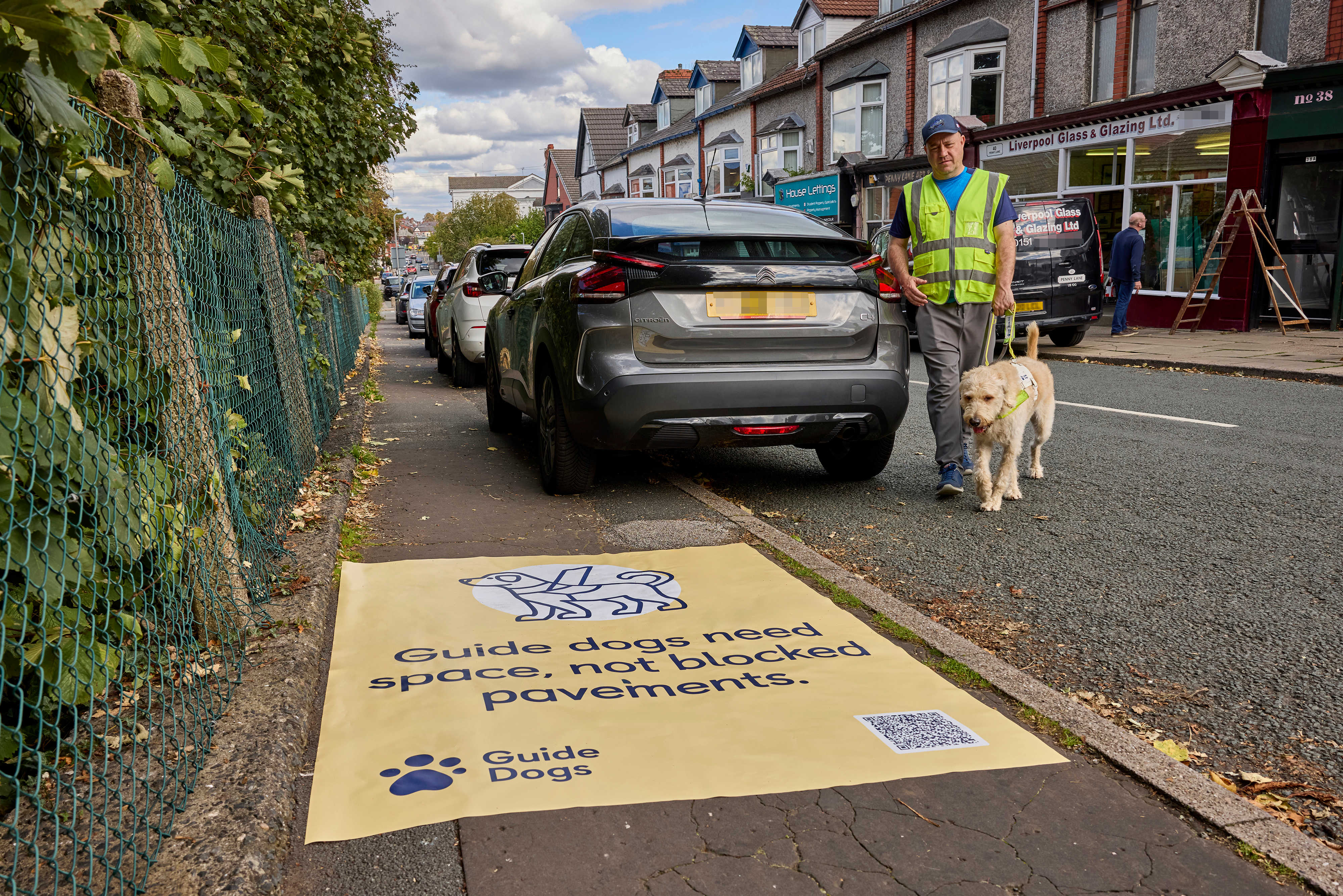 A man and his guide dog walk around the back of a car after being forced into the road because the car is parked on the pavement. In the foreground there is a yellow sticker on the pavement that says "Guide dogs need space, not blocked pavements".