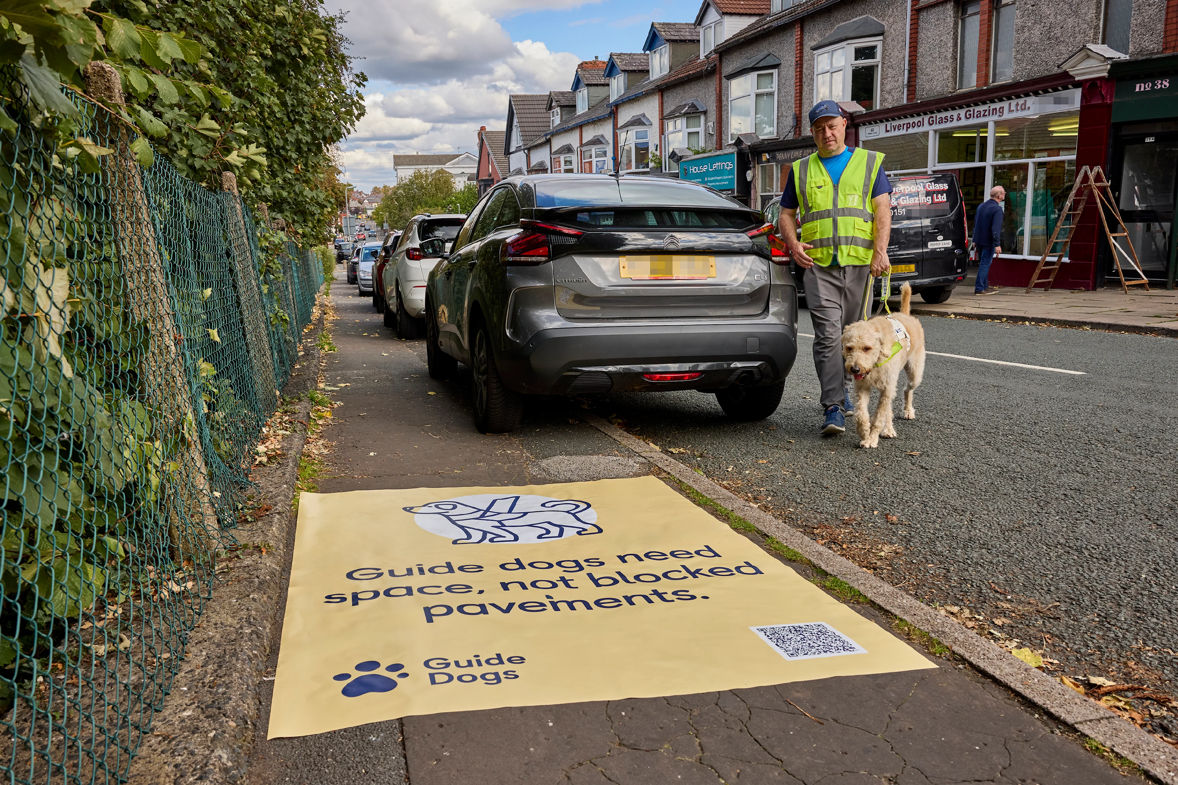 A man and his guide dog walk around the back of a car after being forced into the road because the car is parked on the pavement. In the foreground there is a yellow sticker on the pavement that says "Guide dogs need space, not blocked pavements".