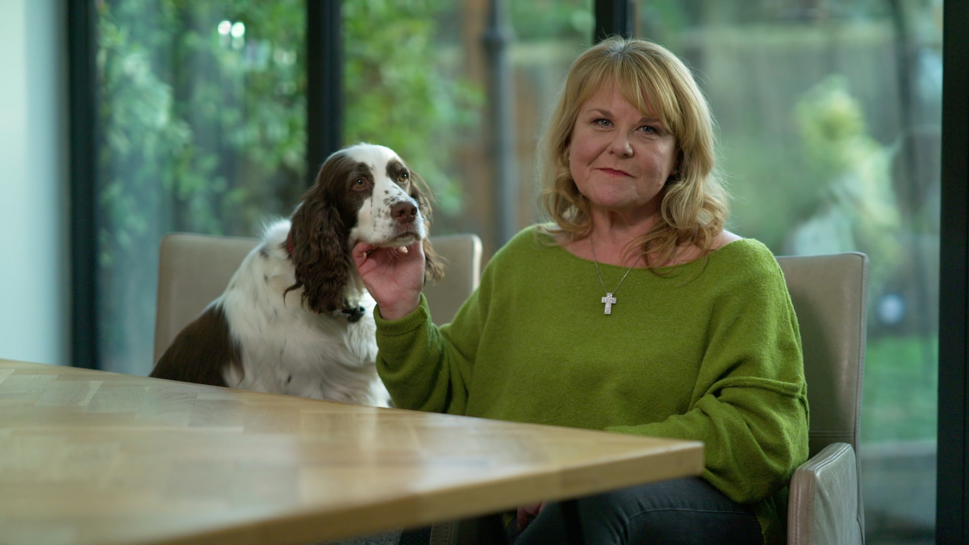 Wendi Peters presents for BBC Lifeline, sitting at a table at home with her pet brown and white springer spaniel on a chair next to her.