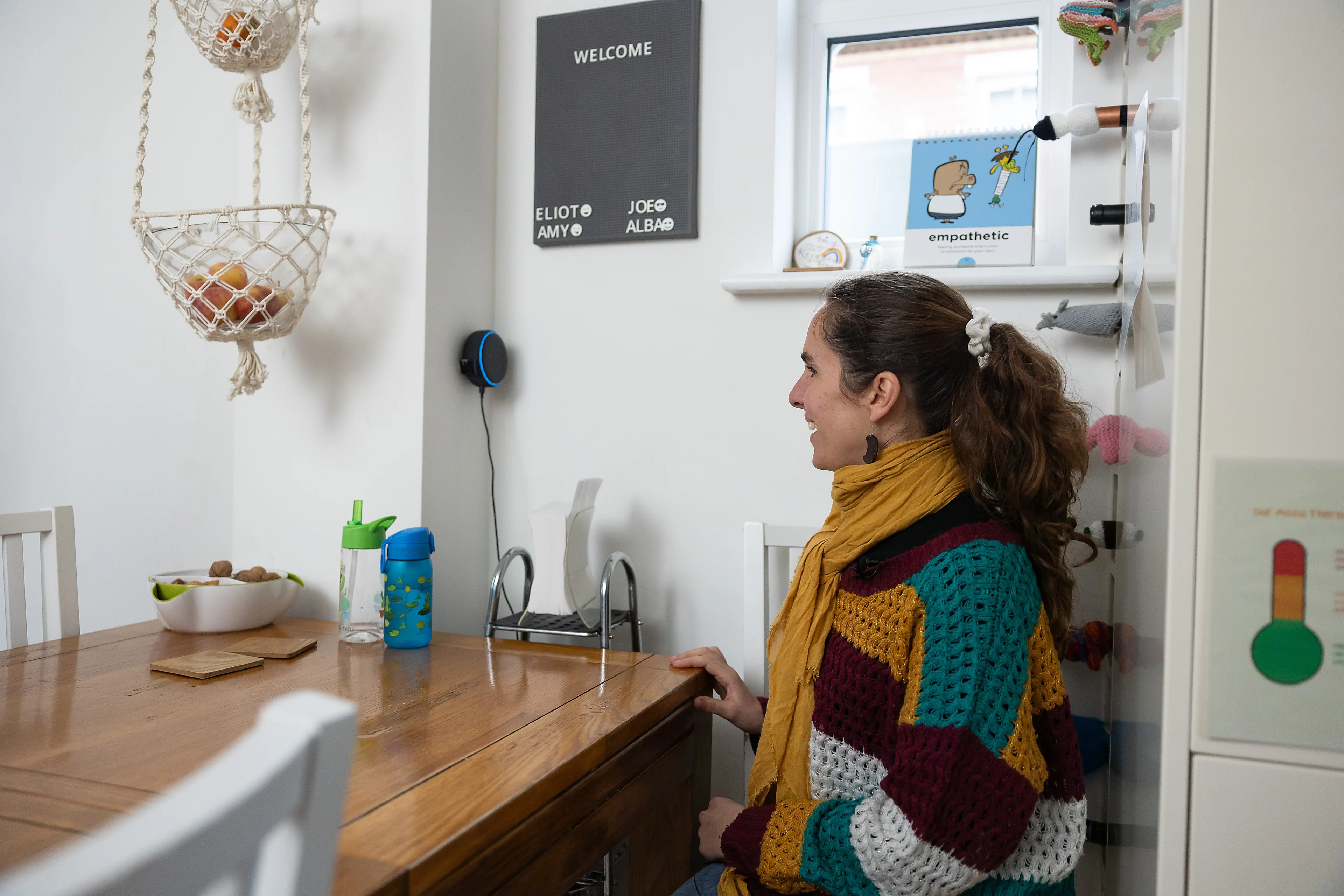 A woman sits at her kitchen table where an Amazon Echo is attached to the wall next to her. She speaks to the Alexa voice assistant. 