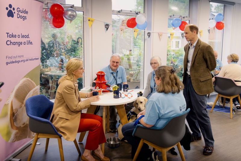 The Duchess of Edinburgh sits at a table with Guide Dogs volunteers while the Duke stands beside them. A guide dog puppy in training sits under the table. The room is decorated with bunting and red, white and blue balloons.