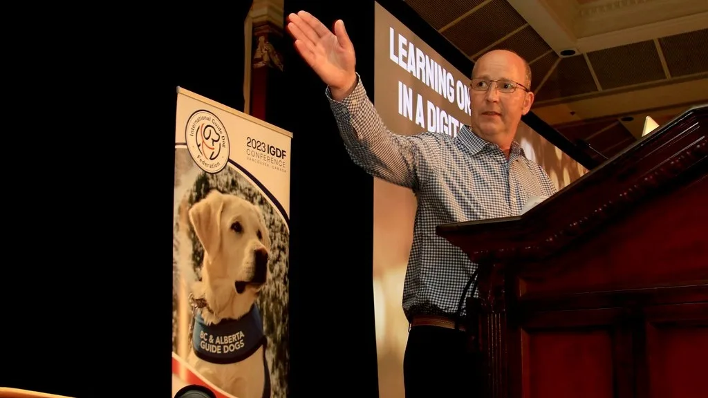 Tim Stafford addresses the IGDF conference at a wooden lectern. A banner with the IGDF logo and a yellow Labrador is behind him. 