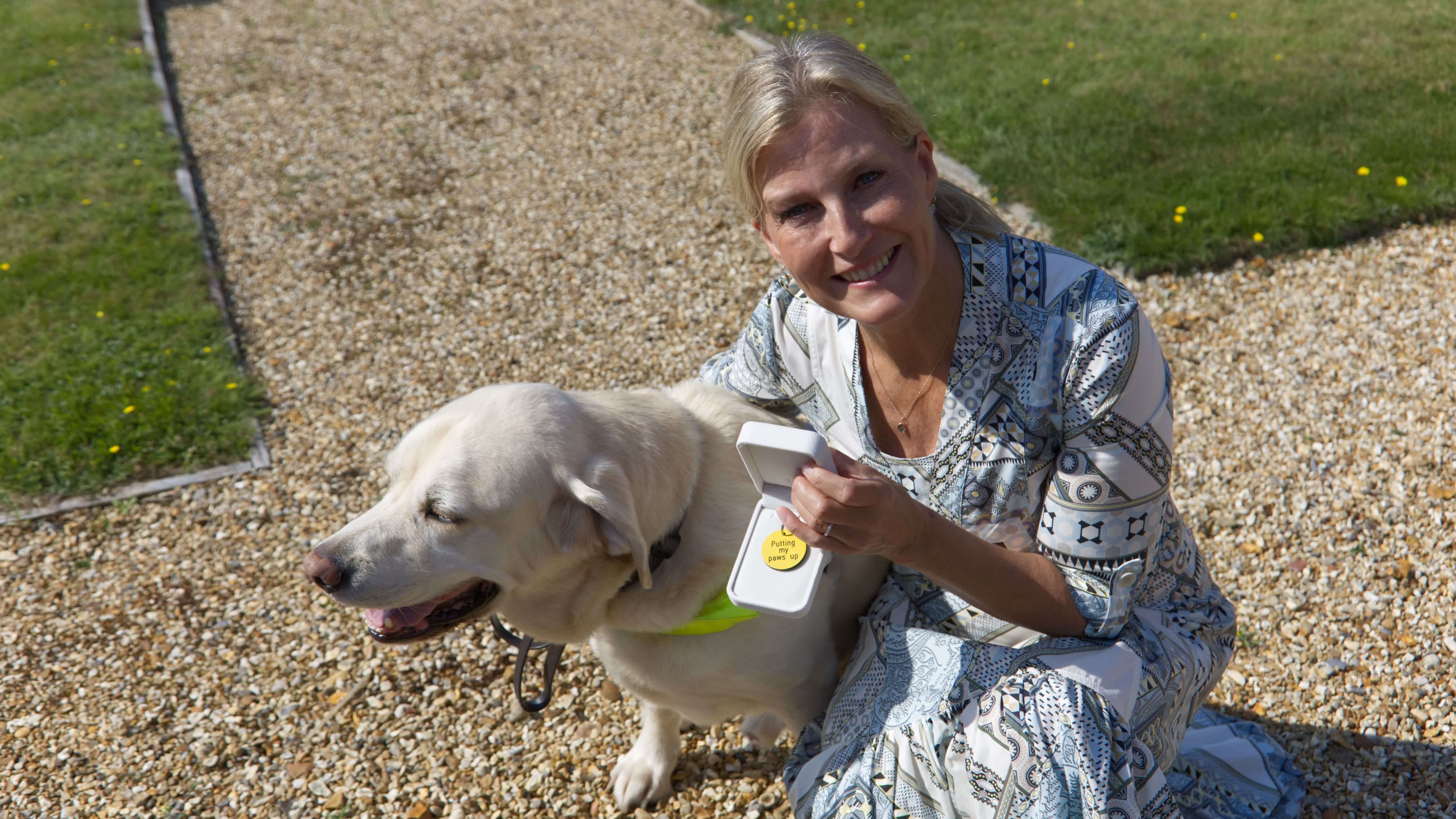 HRH The Duchess of Edinburgh crouches next to retiring guide dog Piper, a yellow Labrador cross golden retriever, while holding a yellow dog tag with the inscription 'Putting my paws up'
