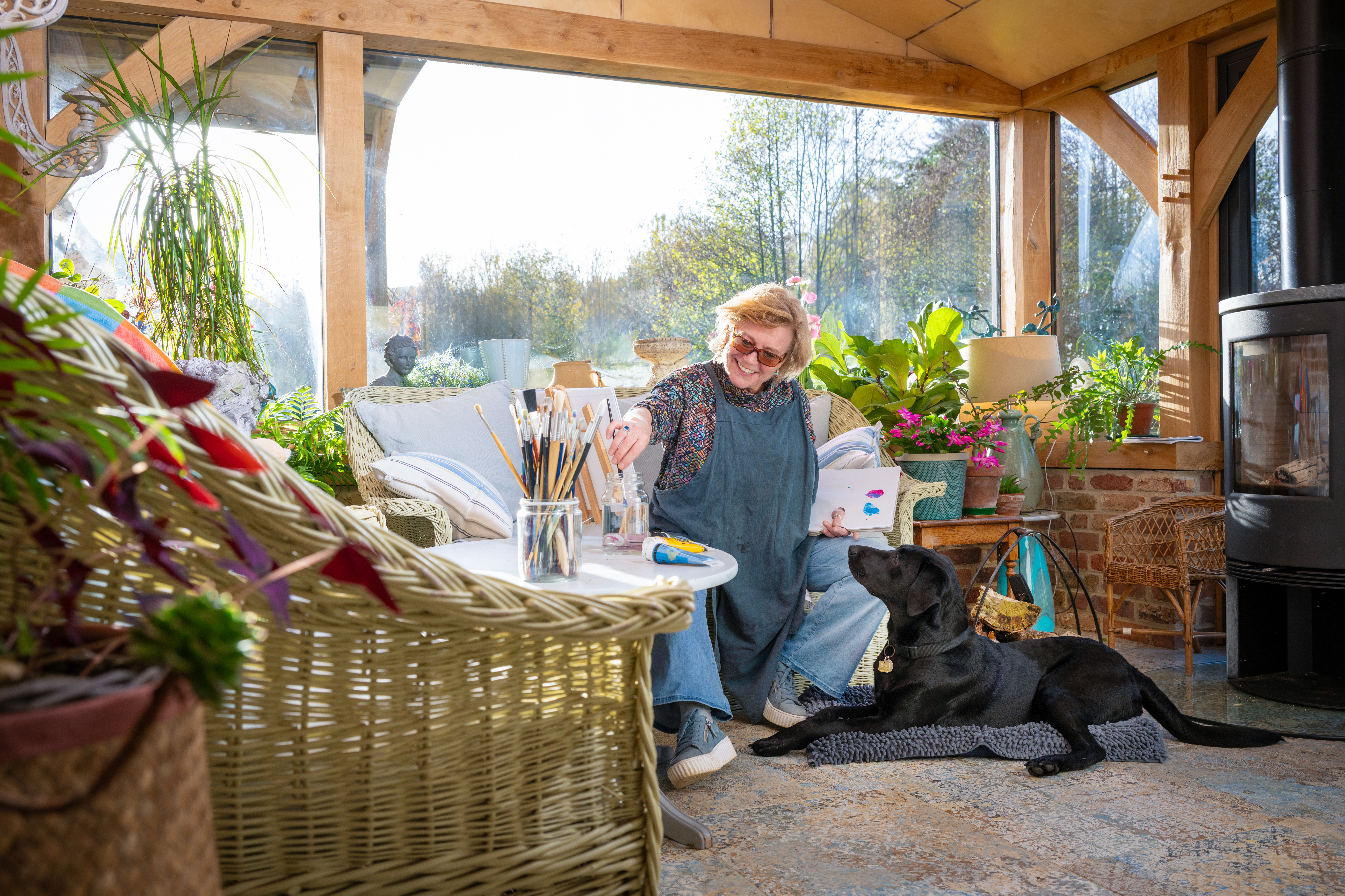 Volunteer puppy raiser Ali sits on a sofa in a bright conservatory and mixes paints with her brushes. A black Labrador guide dog puppy lies on the floor besides Ali and looks up at her. 