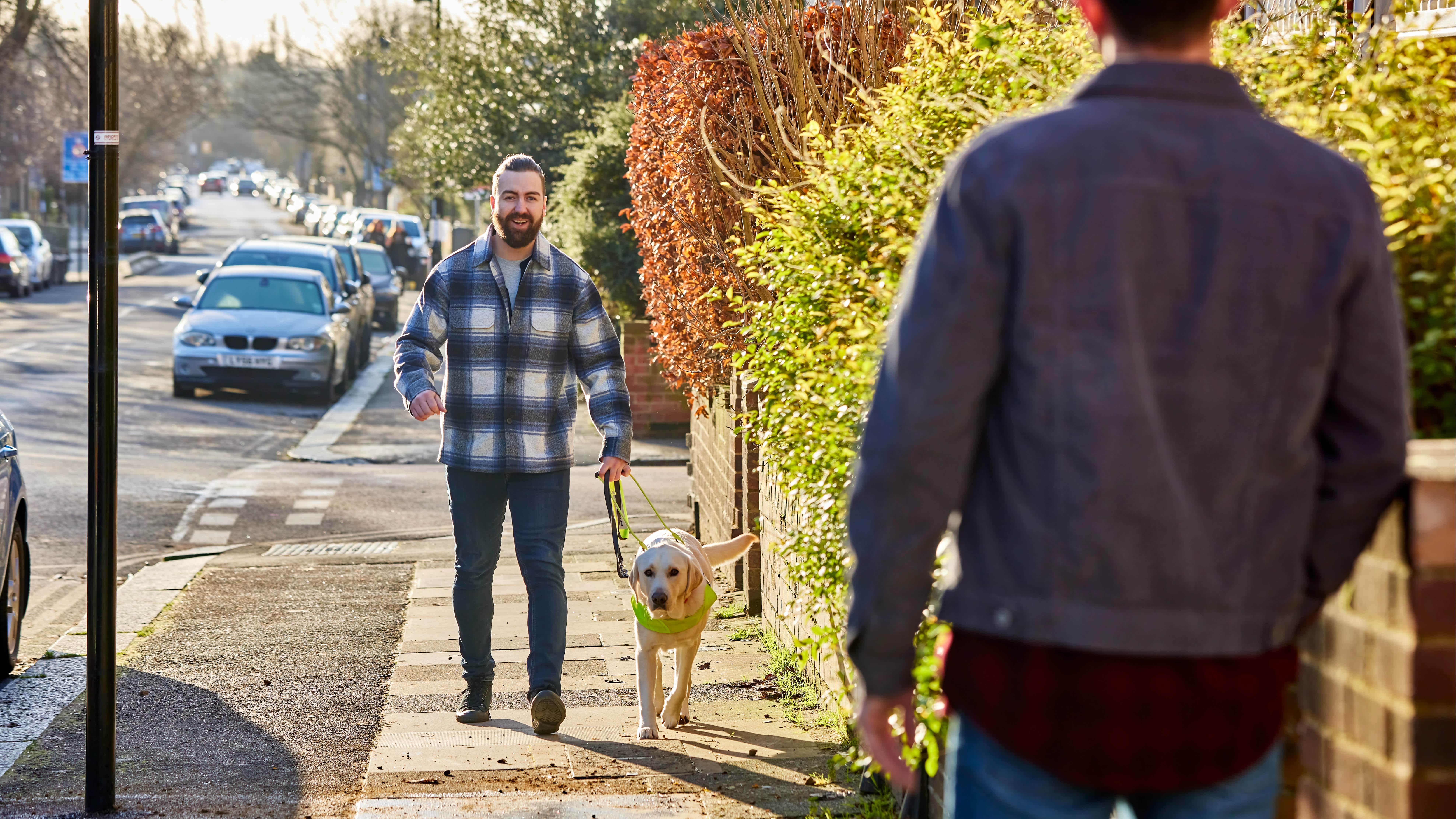 In the foreground we see the back of a man wearing a denim jacket. He is facing his brother, a man with a beard wearing a checked shirt, who is walking up the road with his guide dog, a yellow labrador in a Guide Dogs harness.