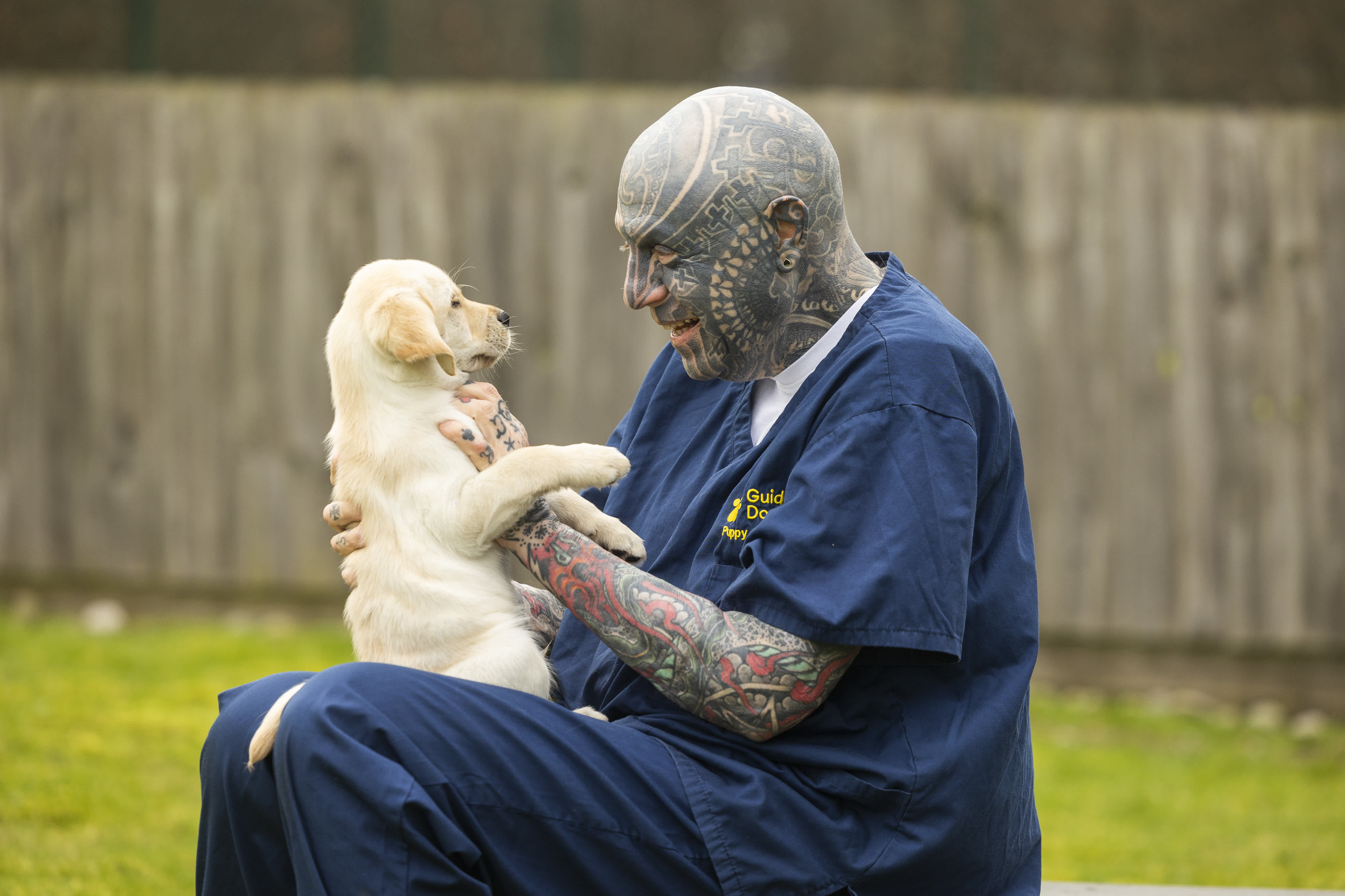 A man with lots of blue coloured tattoos covering his head and arms sits wearing Guide Dogs branded blue scrubs. He is holding a young yellow Labrador guide dog puppy in his lap. 