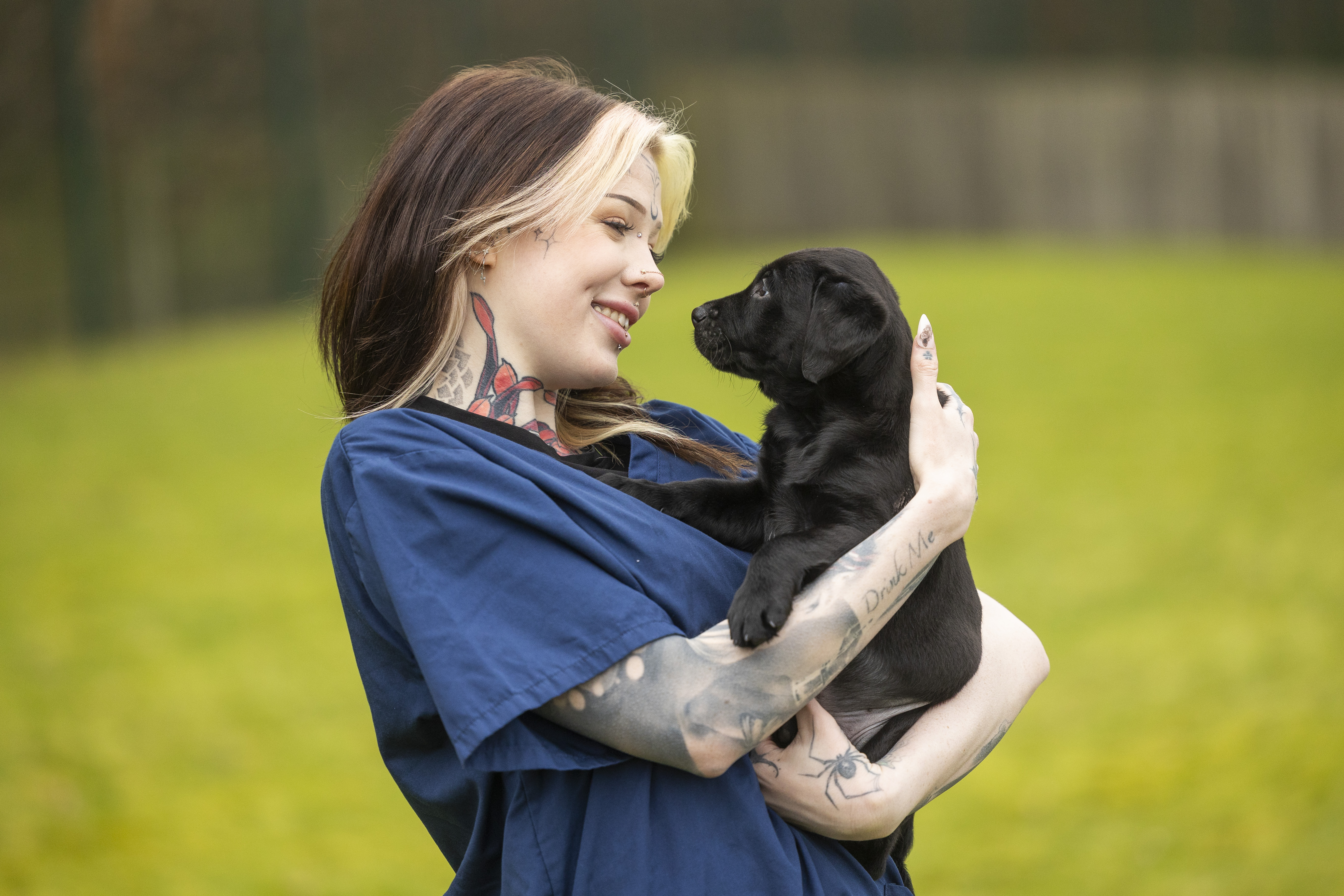 A woman with blonde and brown hair and facial tattoos and piercings wearing Guide Dogs branded navy blue scrubs is holding and smiling at a young black Labrador guide dog puppy. 